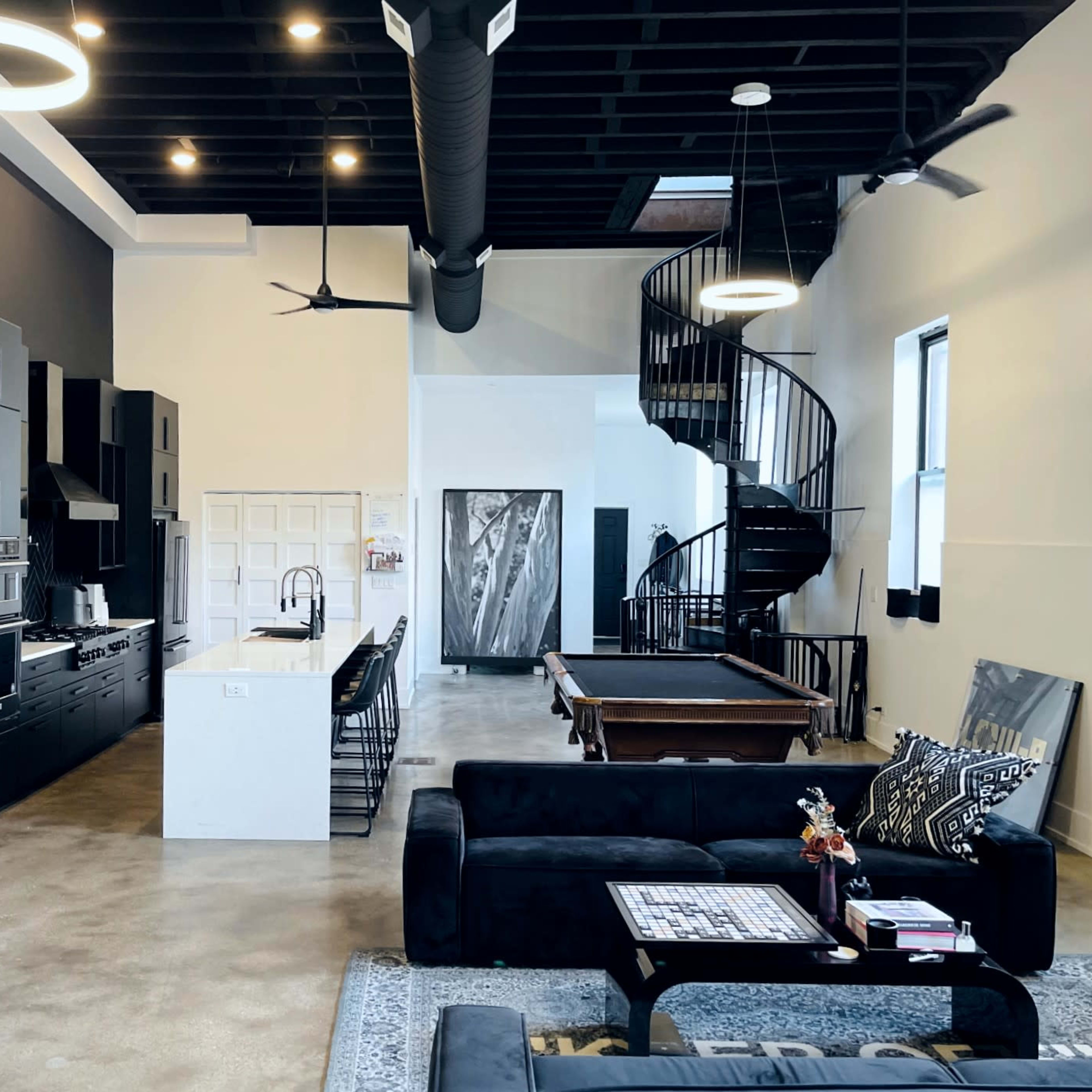 A modern open-concept living space features a kitchen with black cabinetry, a spiral staircase, and a pool table adjacent to a seating area with a black sofa and decorative elements.