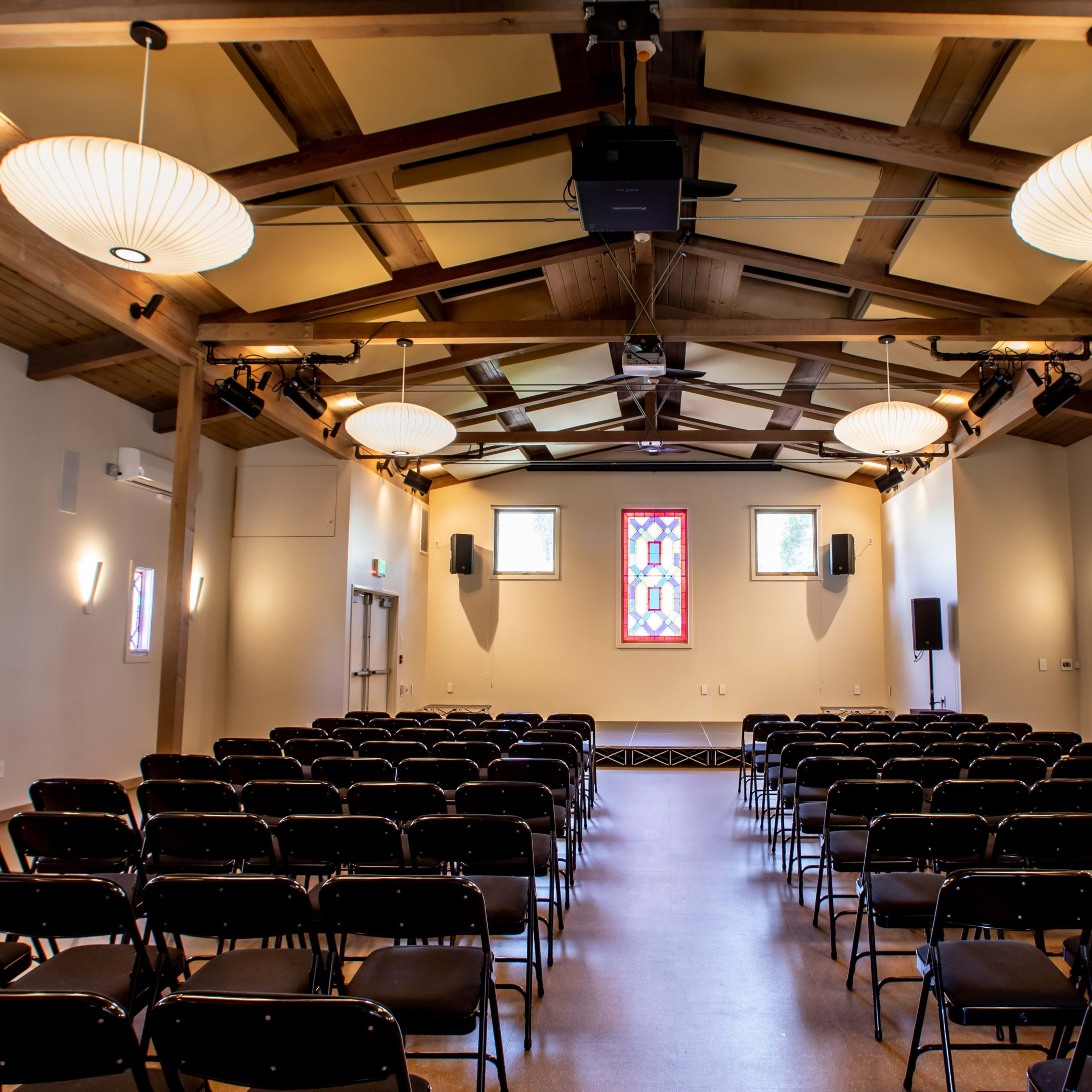 A spacious indoor hall with rows of black chairs facing a stained glass window and a wall-mounted screen.