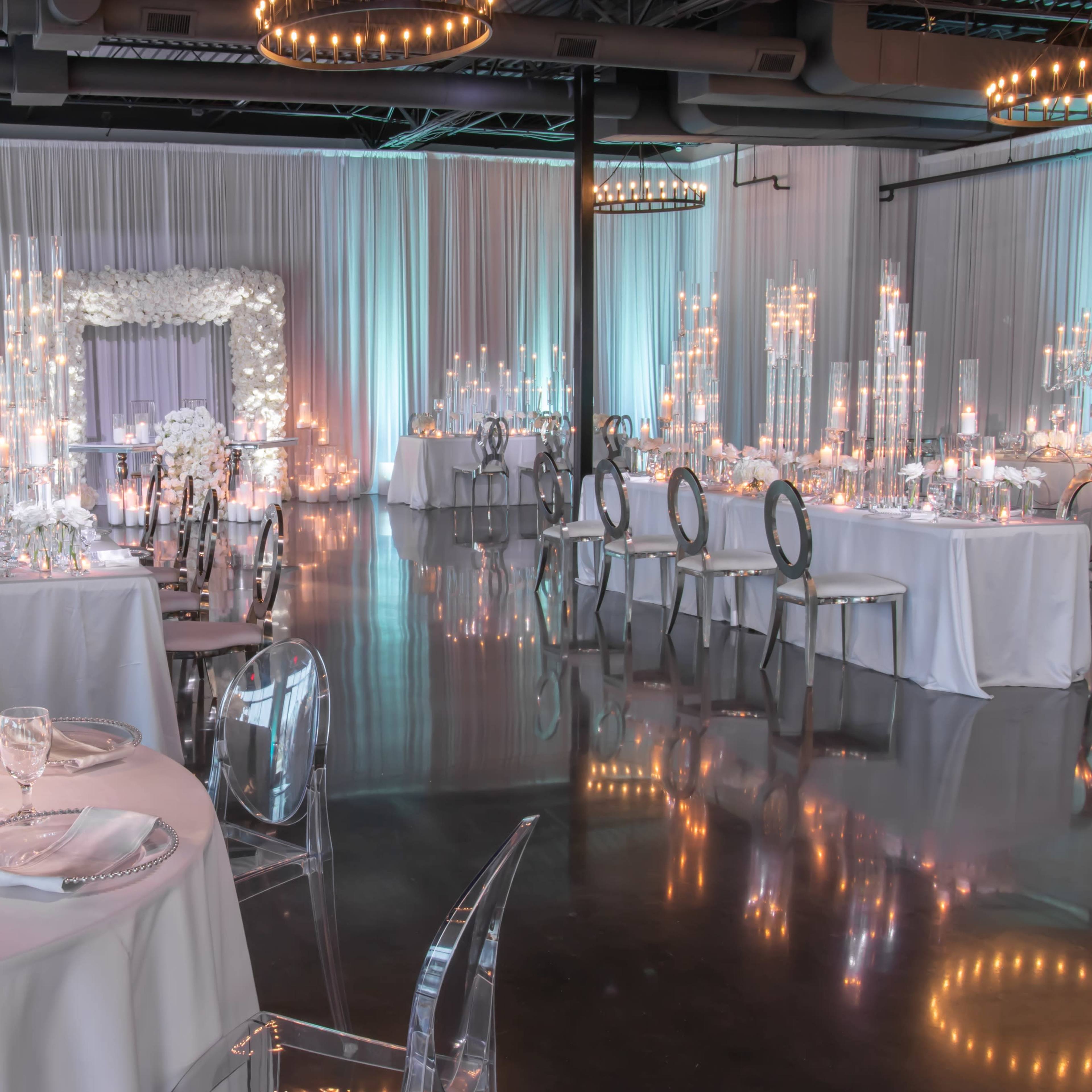 An elegantly decorated event space with tables set for a formal gathering, featuring white tablecloths, floral centerpieces, and ambient candlelight.