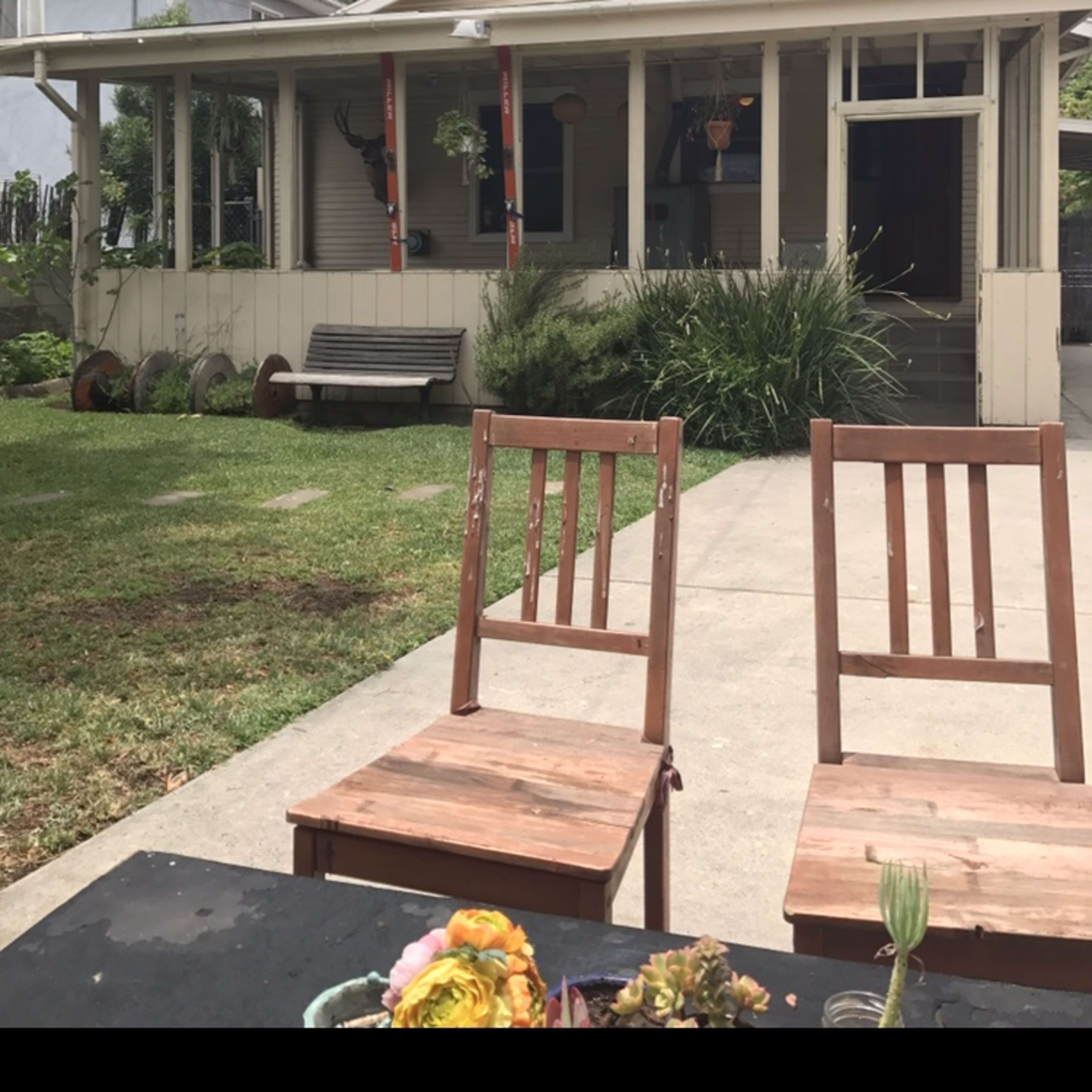 A wooden table with two empty chairs is positioned in front of a house featuring a porch and surrounding greenery.
