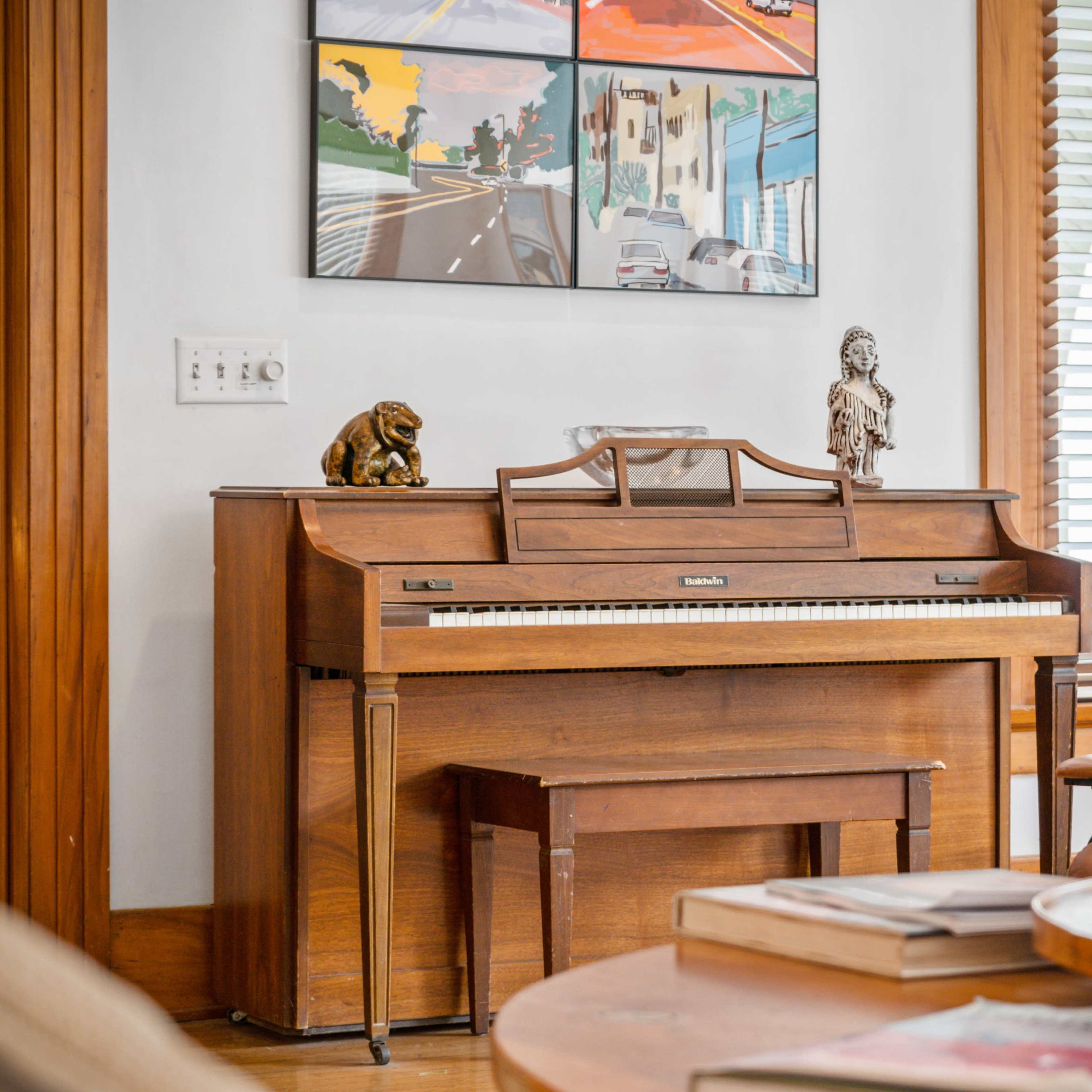 A wooden piano stands against a wall, accompanied by a small bench, framed art on the wall, and various decorative items on a coffee table.