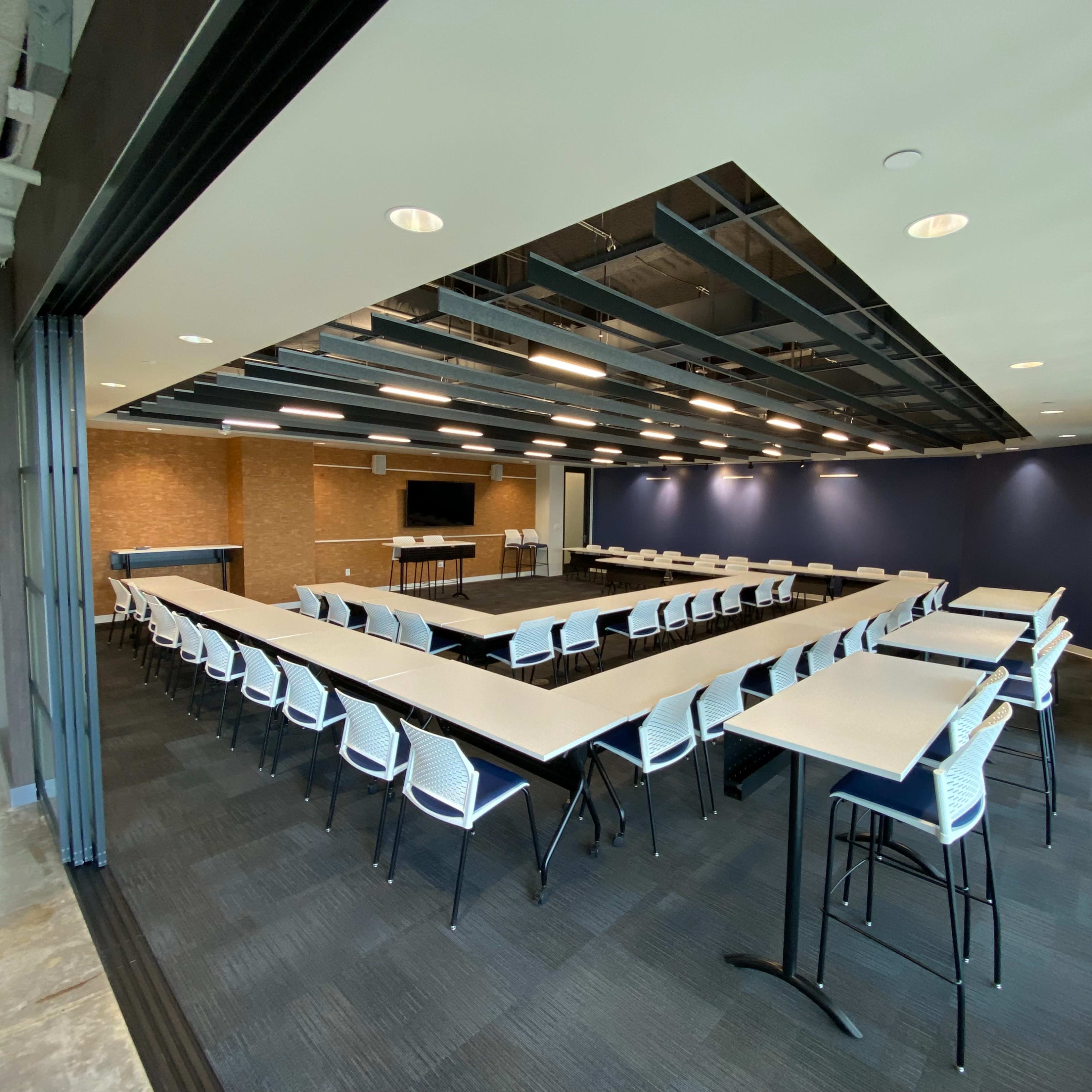 A modern conference room arranged in a U-shape with white chairs and tables, featuring a large screen at one end and dark blue walls.