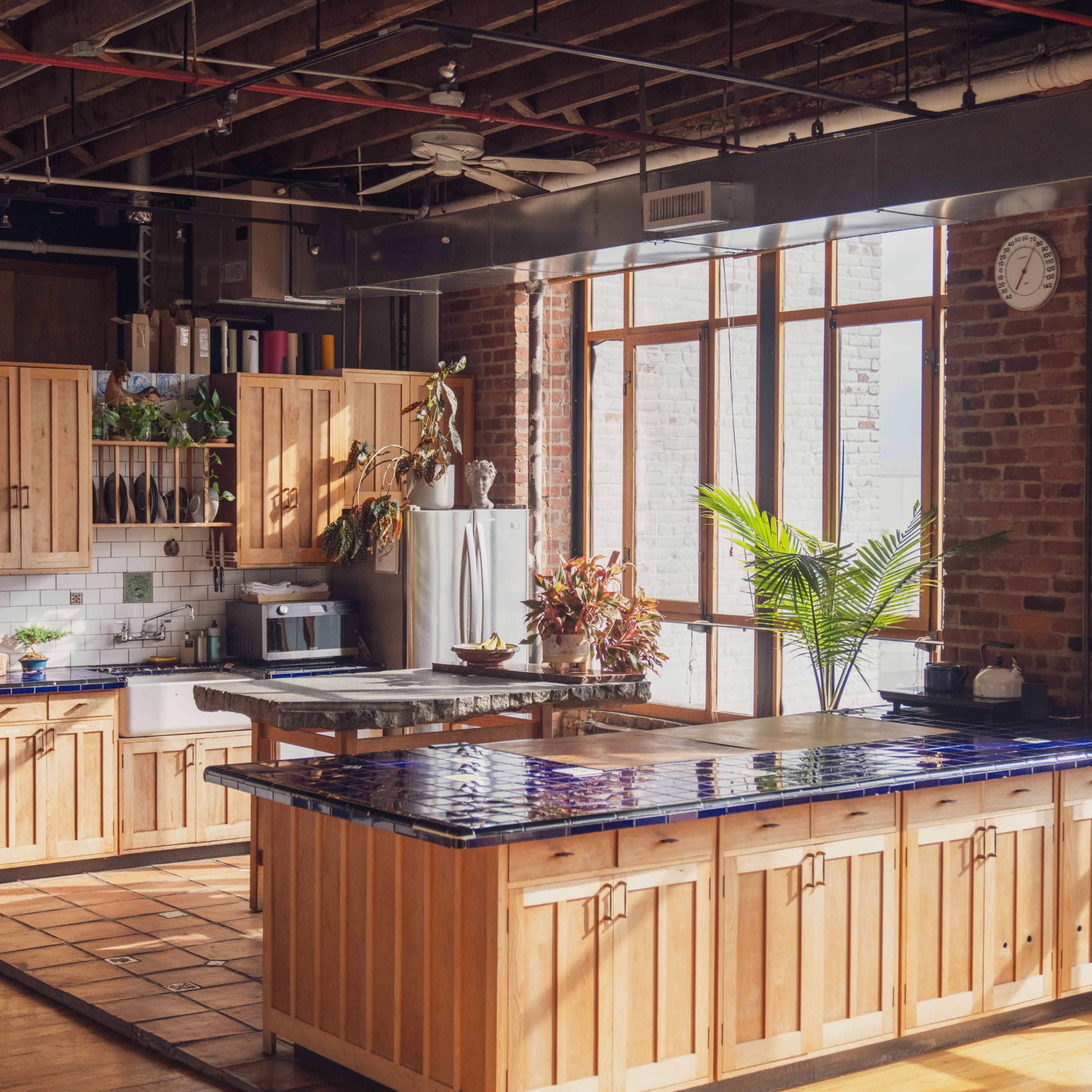 The kitchen features wooden cabinets, a tiled island, and large windows that bring in natural light, all set against exposed brick walls.