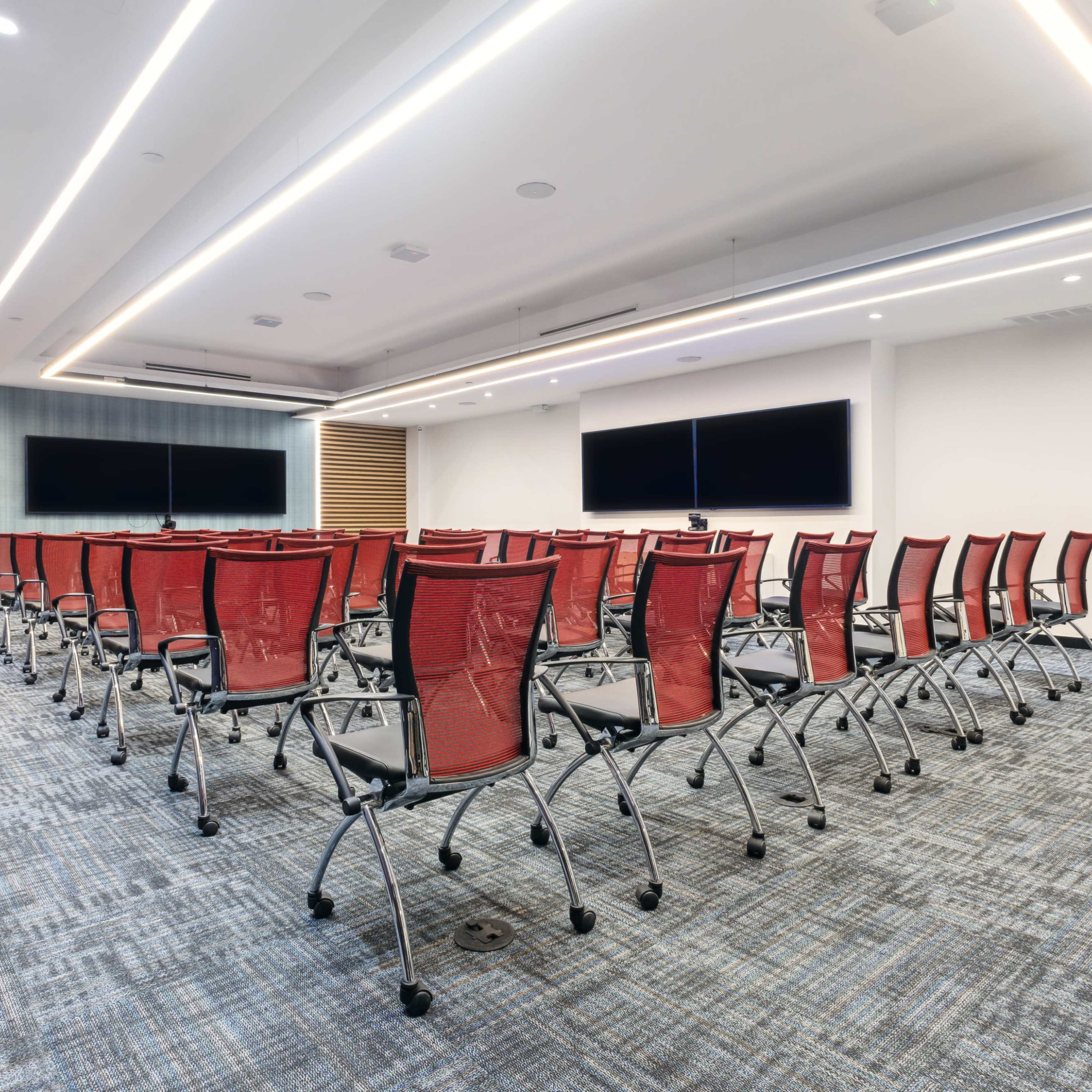 A modern conference room arranged with rows of red mesh chairs facing two large screens on the front wall.