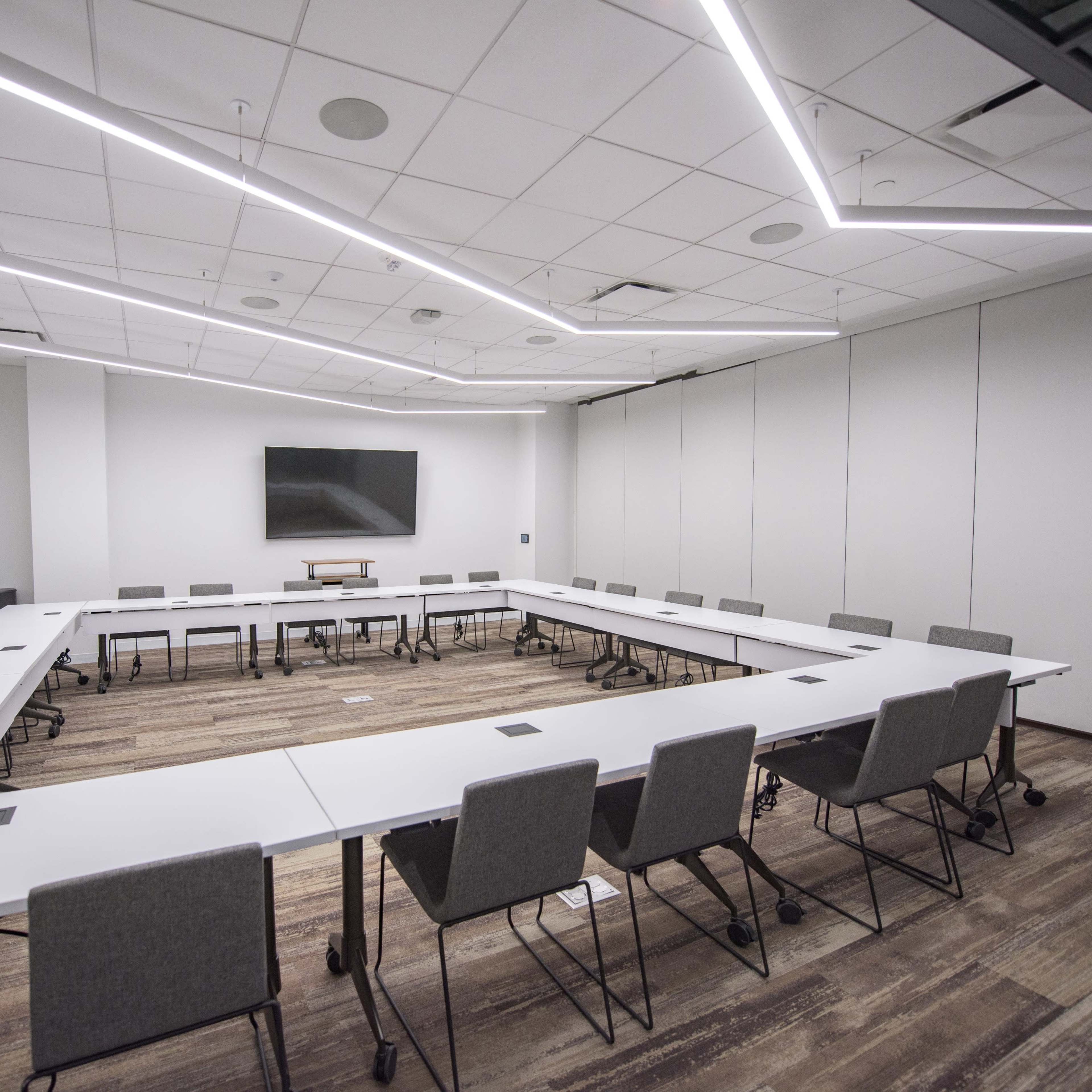A modern conference room features a large rectangular table surrounded by gray chairs, with a wall-mounted screen and recessed lighting.