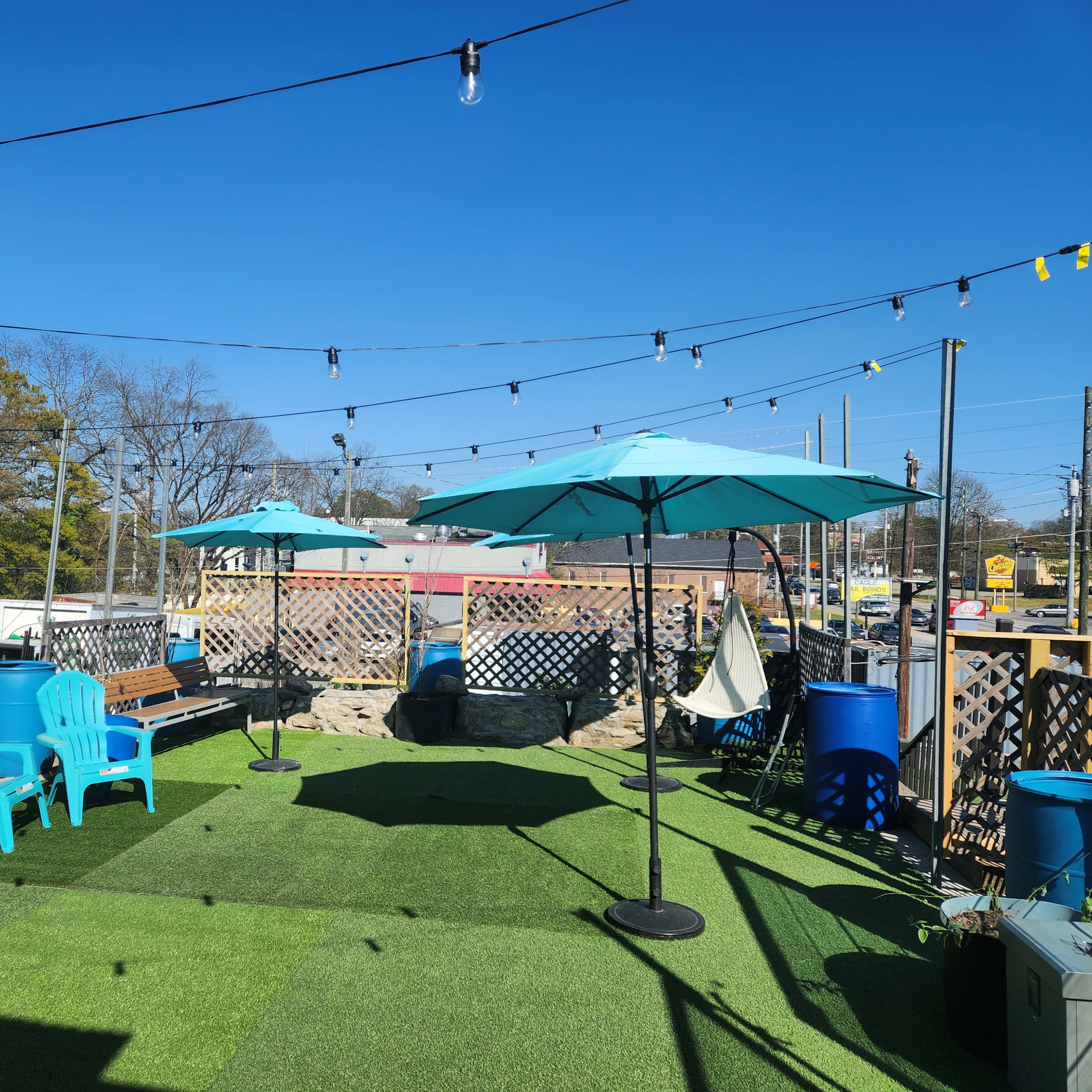 A rooftop area features turquoise umbrellas, blue chairs, and string lights over a green artificial grass surface.