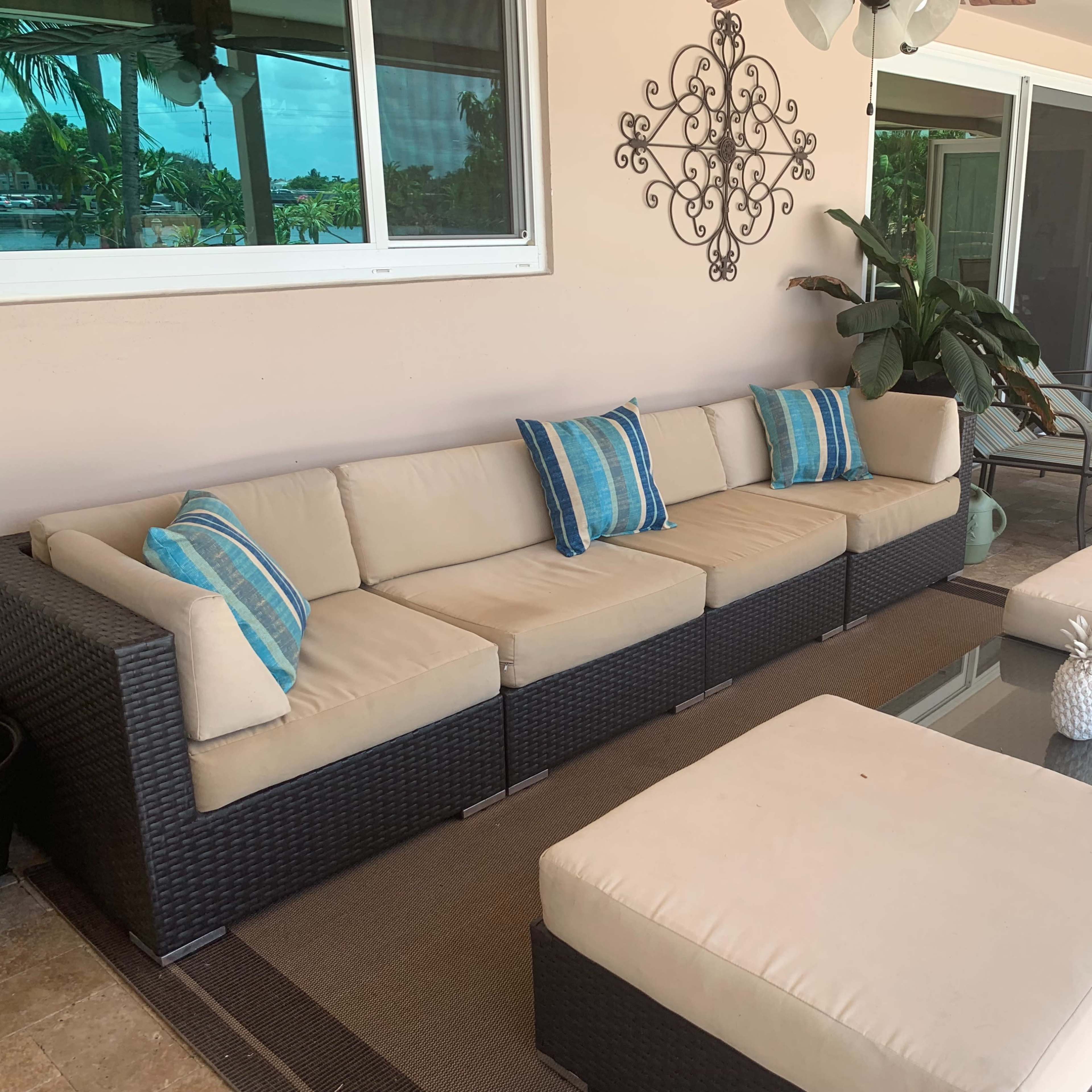 A spacious outdoor seating area features a light-colored sectional sofa with decorative pillows, a glass coffee table, and surrounding greenery.