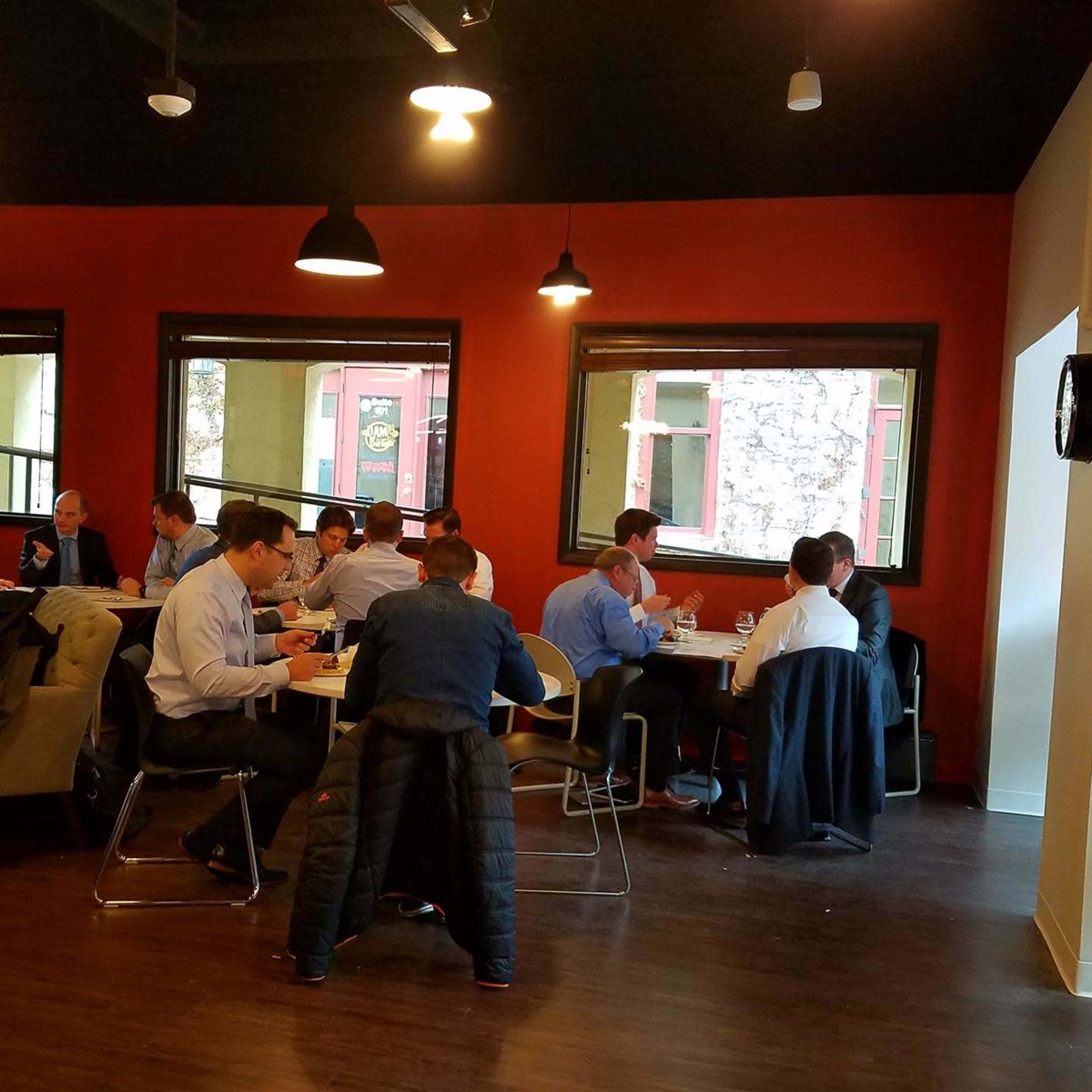 A group of people in business attire sits at tables in a restaurant with large windows and a mix of dark and light decor.
