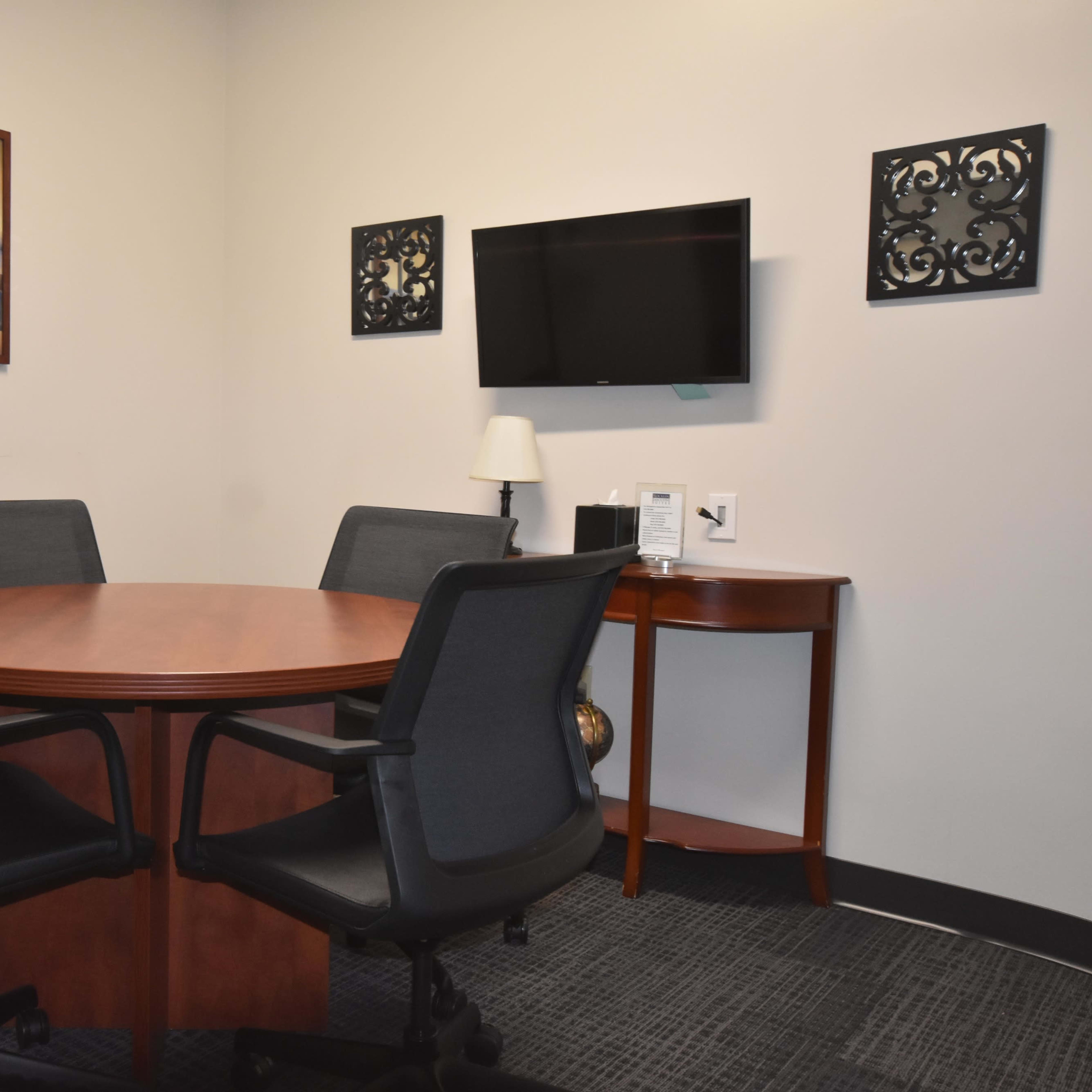 A small conference room features a circular wooden table surrounded by four black chairs, with wall artwork and a mounted television.
