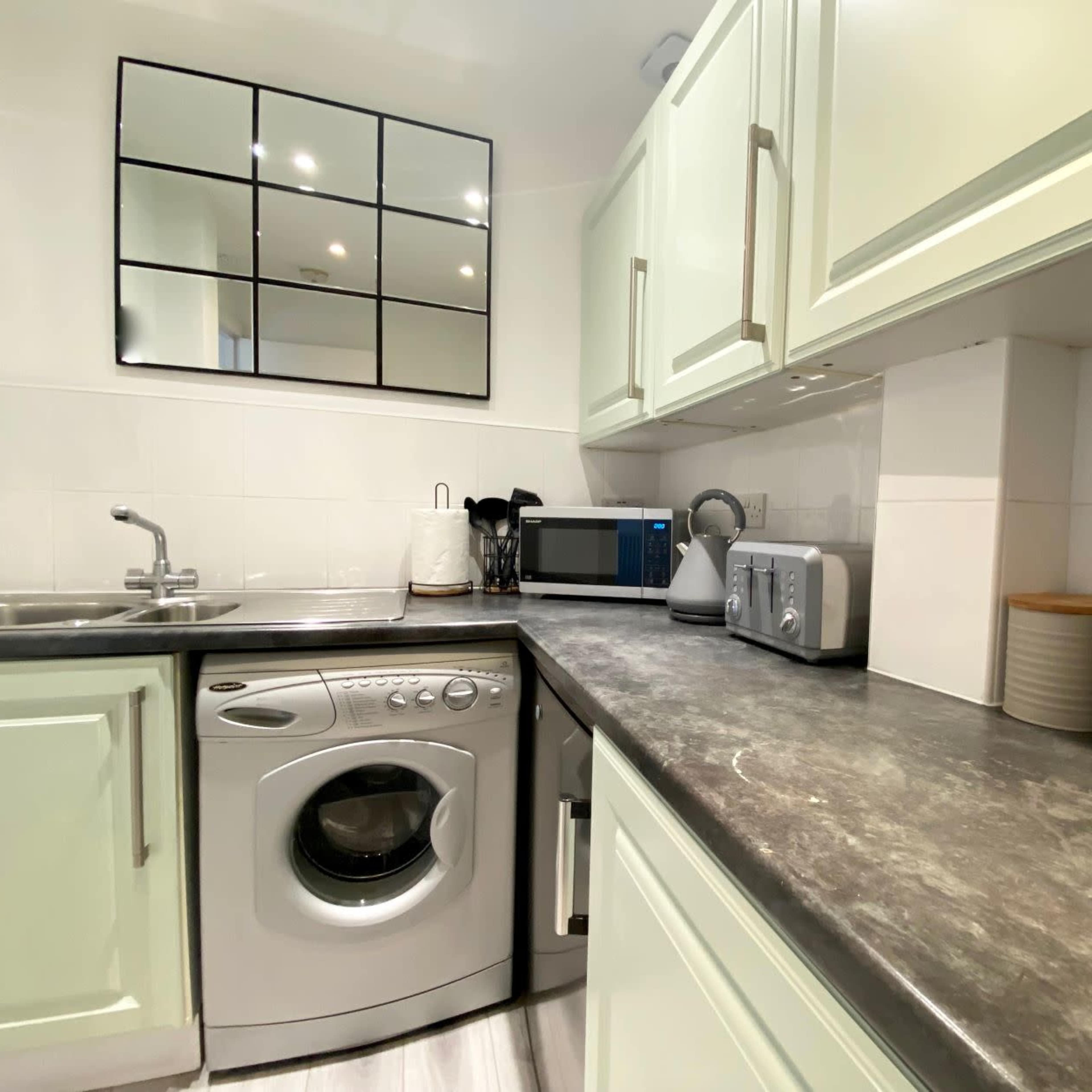 A compact kitchen features a washing machine, sink, microwave, kettle, toaster, and neatly arranged canisters on a countertop.