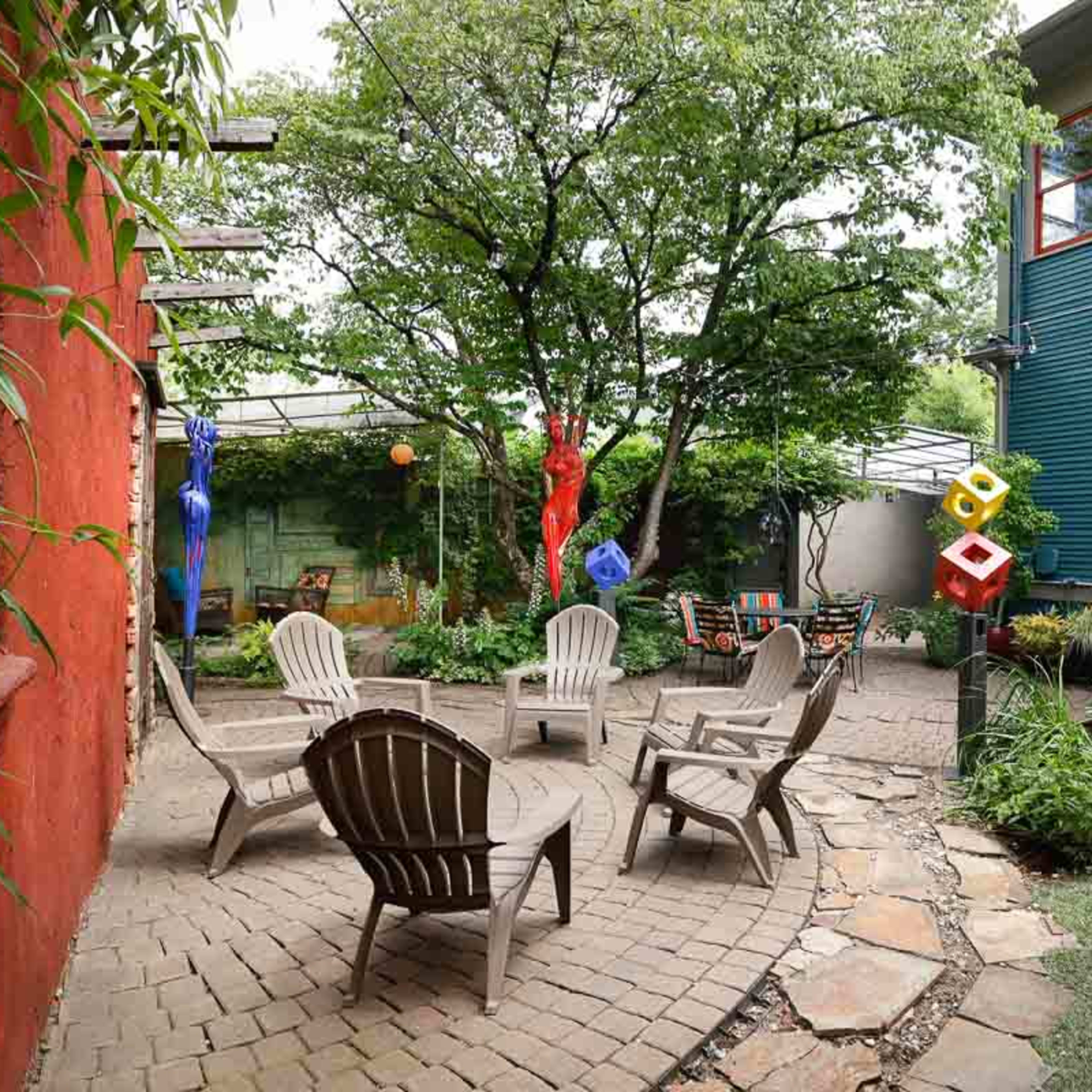 A landscaped courtyard with a circular arrangement of wooden chairs around a stone patio, surrounded by greenery and colorful decorative items.