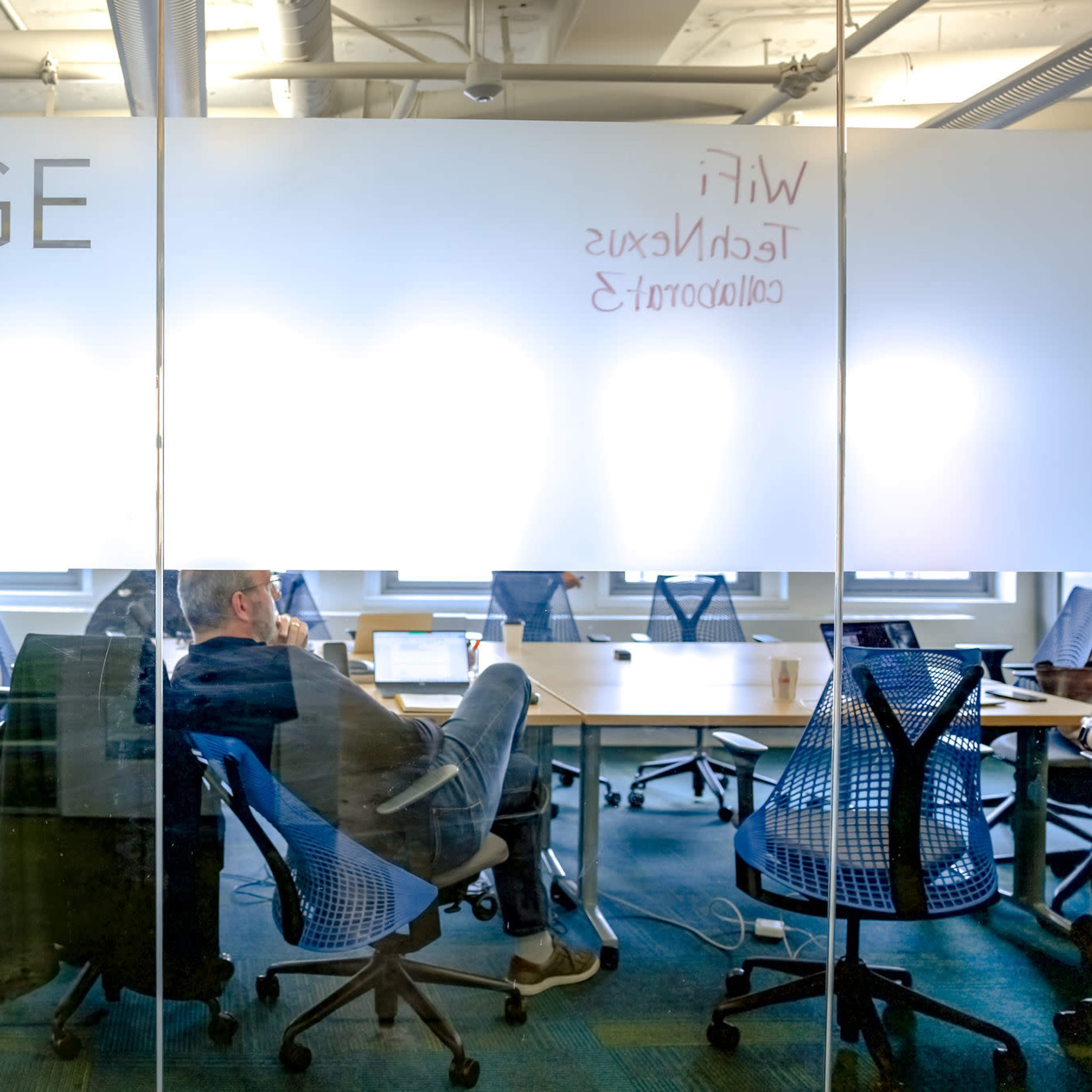 A person sits in a chair with their feet on a table in a glass-walled office labeled "BABBAGE," with scattered chairs and a whiteboard visible in the background.