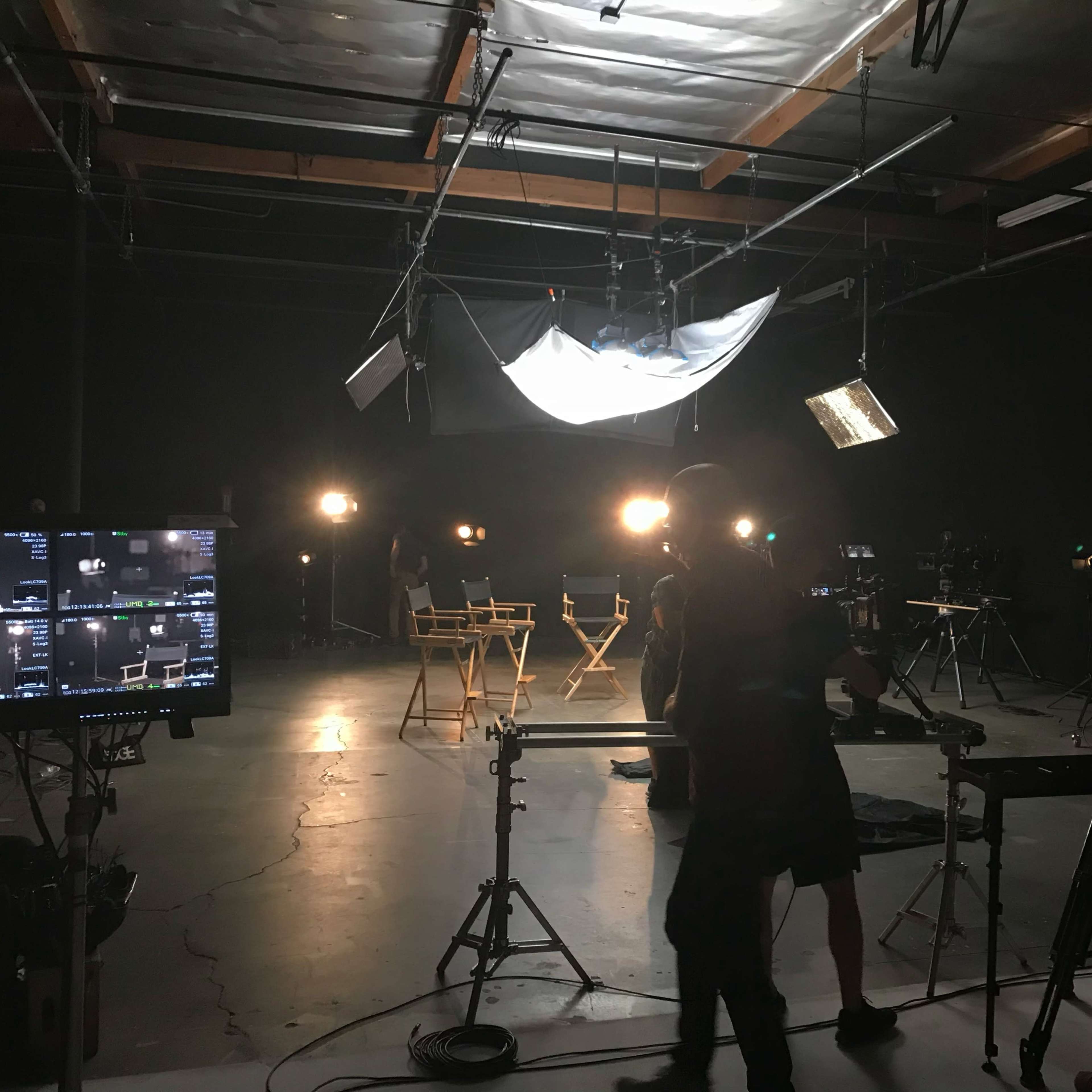 A film studio setup features multiple cameras, lighting equipment, and two director's chairs positioned in front of a black backdrop.