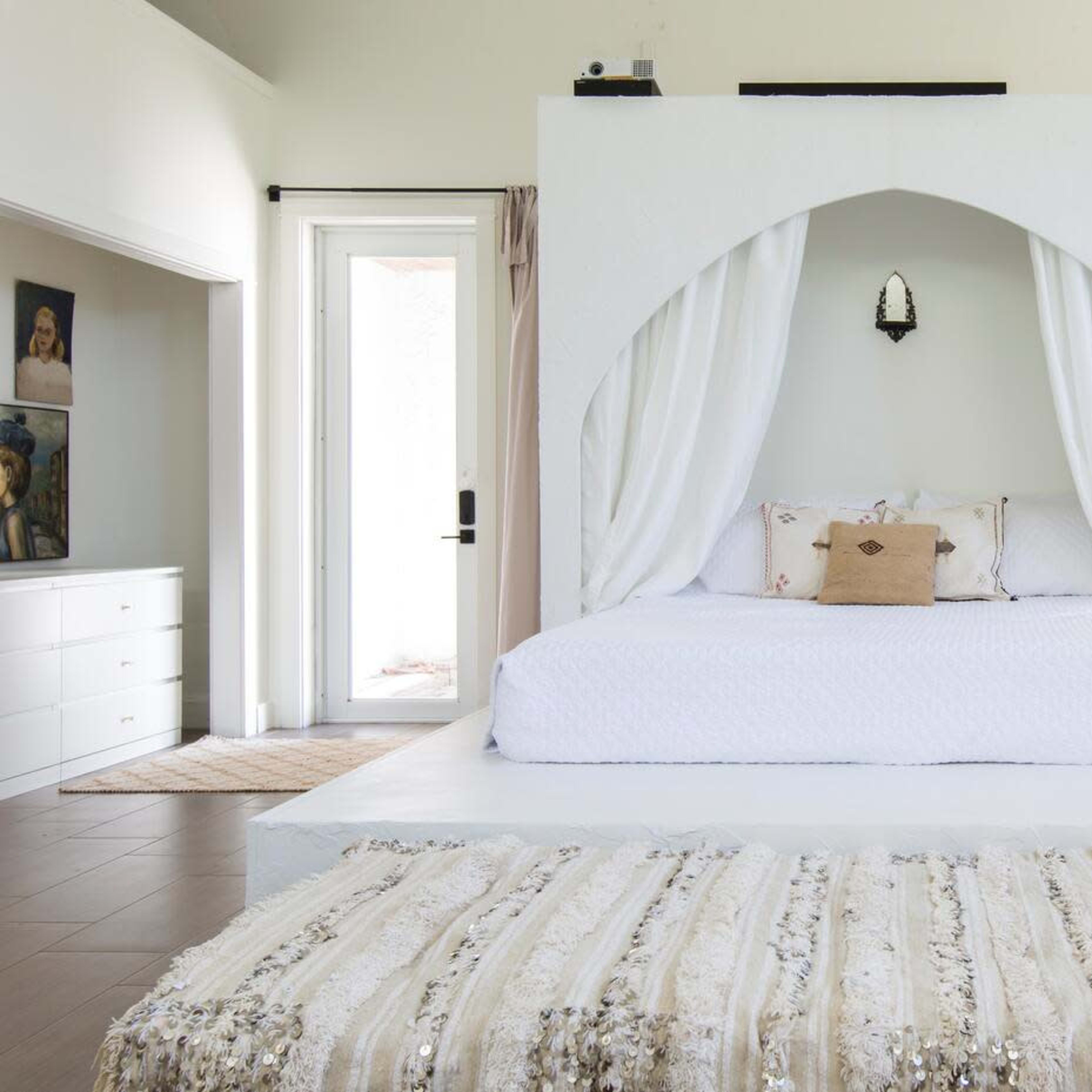A modern bedroom with a raised, white four-poster bed draped in curtains, positioned on a light-colored floor with a decorative rug in front.