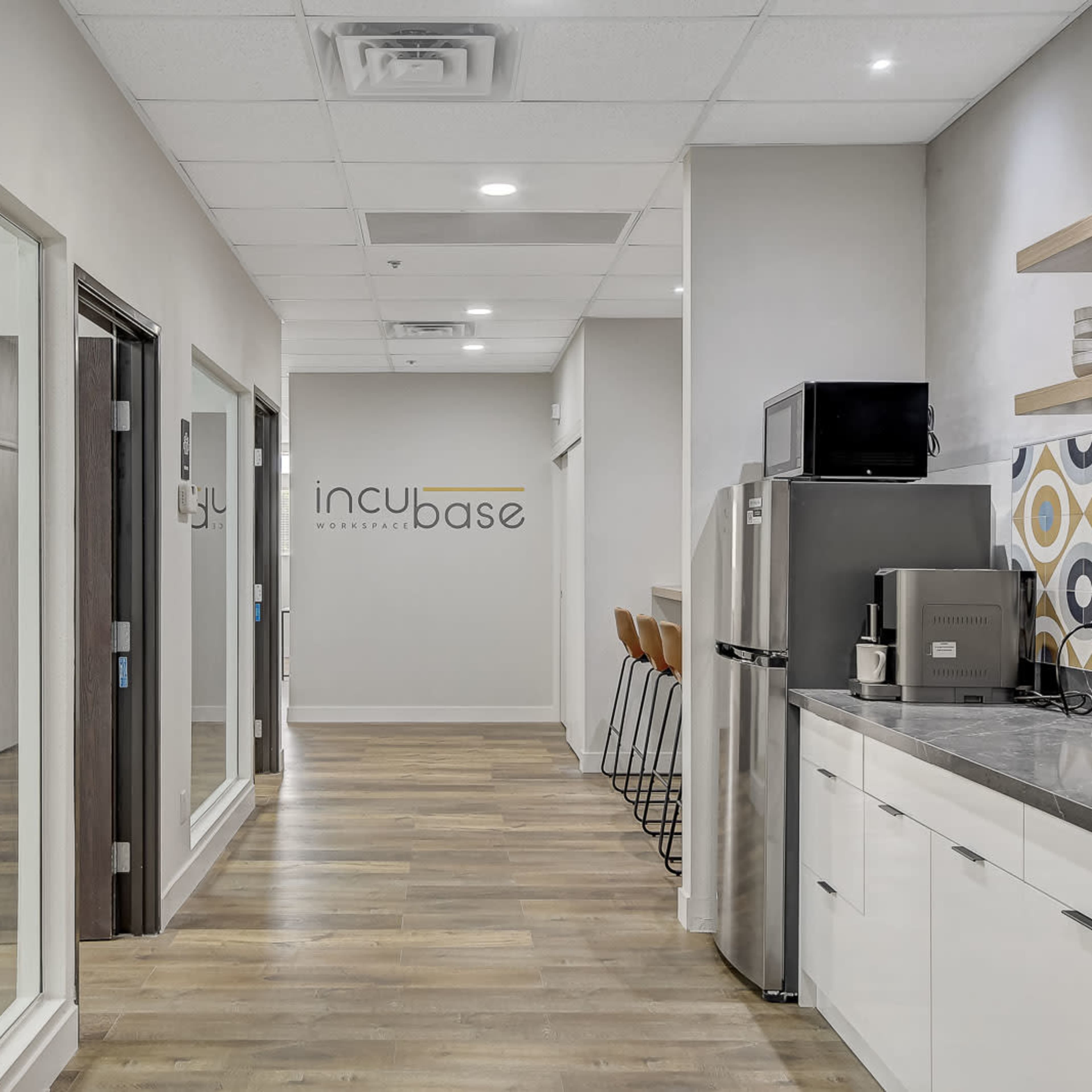 A modern office hallway leading to a kitchenette and several glass-walled offices, with a logo reading "incubase" on the wall.