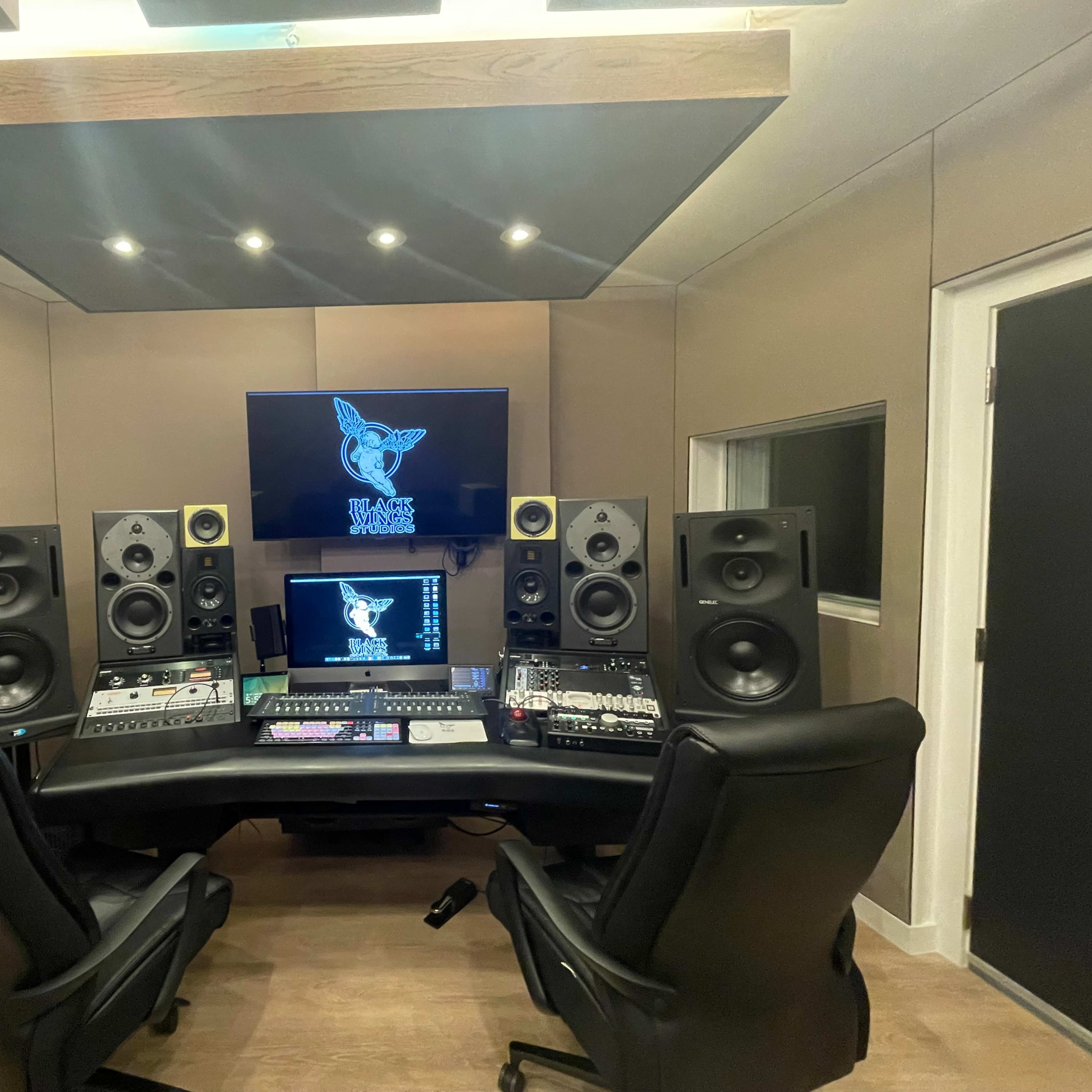 A modern music production studio featuring two black chairs, a large monitor displaying a logo, and multiple speakers arranged around a mixing console.