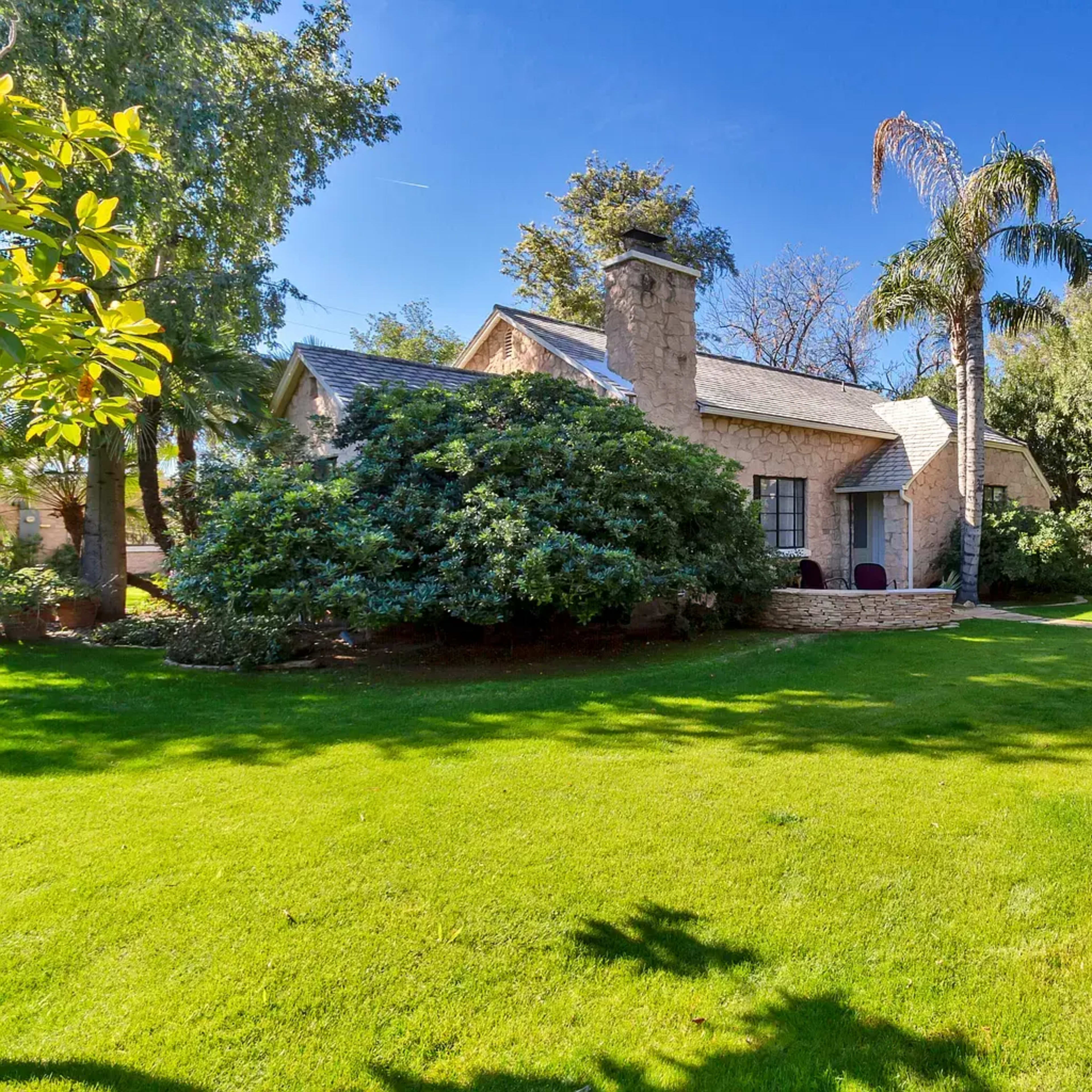 A small house with a stone chimney is surrounded by a well-maintained lawn and several trees.