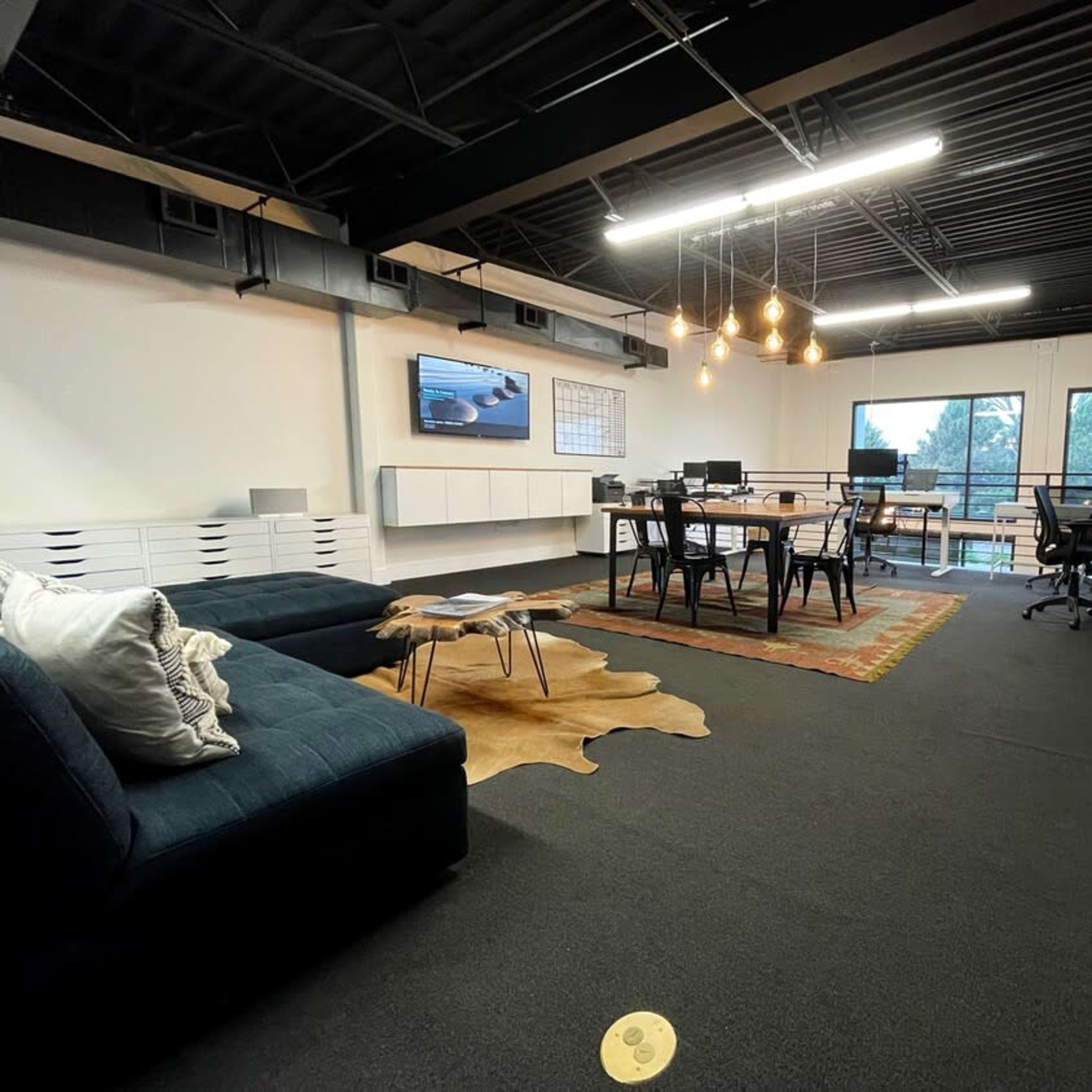 A modern office space with a seating area, a dining table, and large windows overlooking greenery.