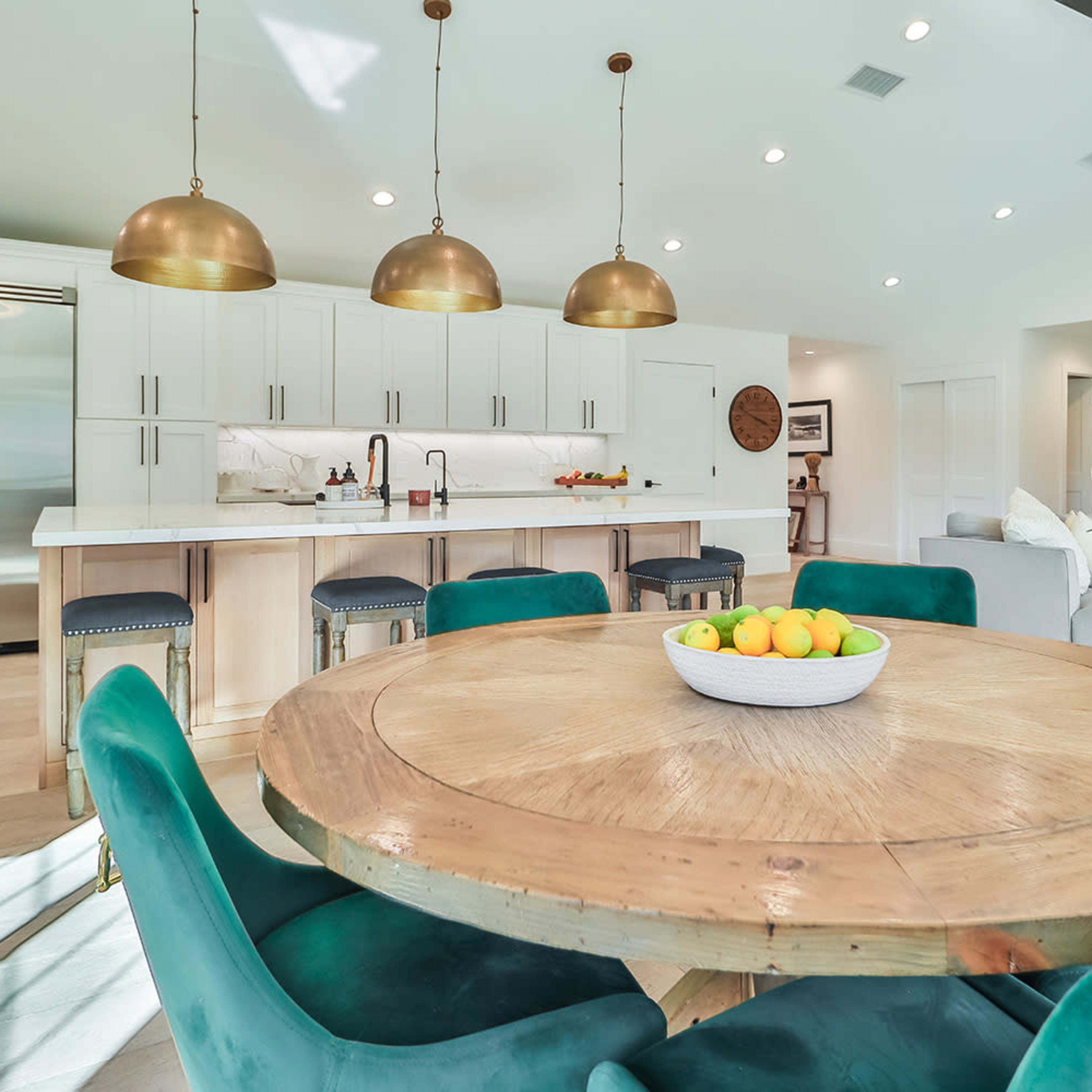 A modern kitchen with a large round wooden dining table surrounded by green chairs, featuring pendant lights and stainless steel appliances in the background.