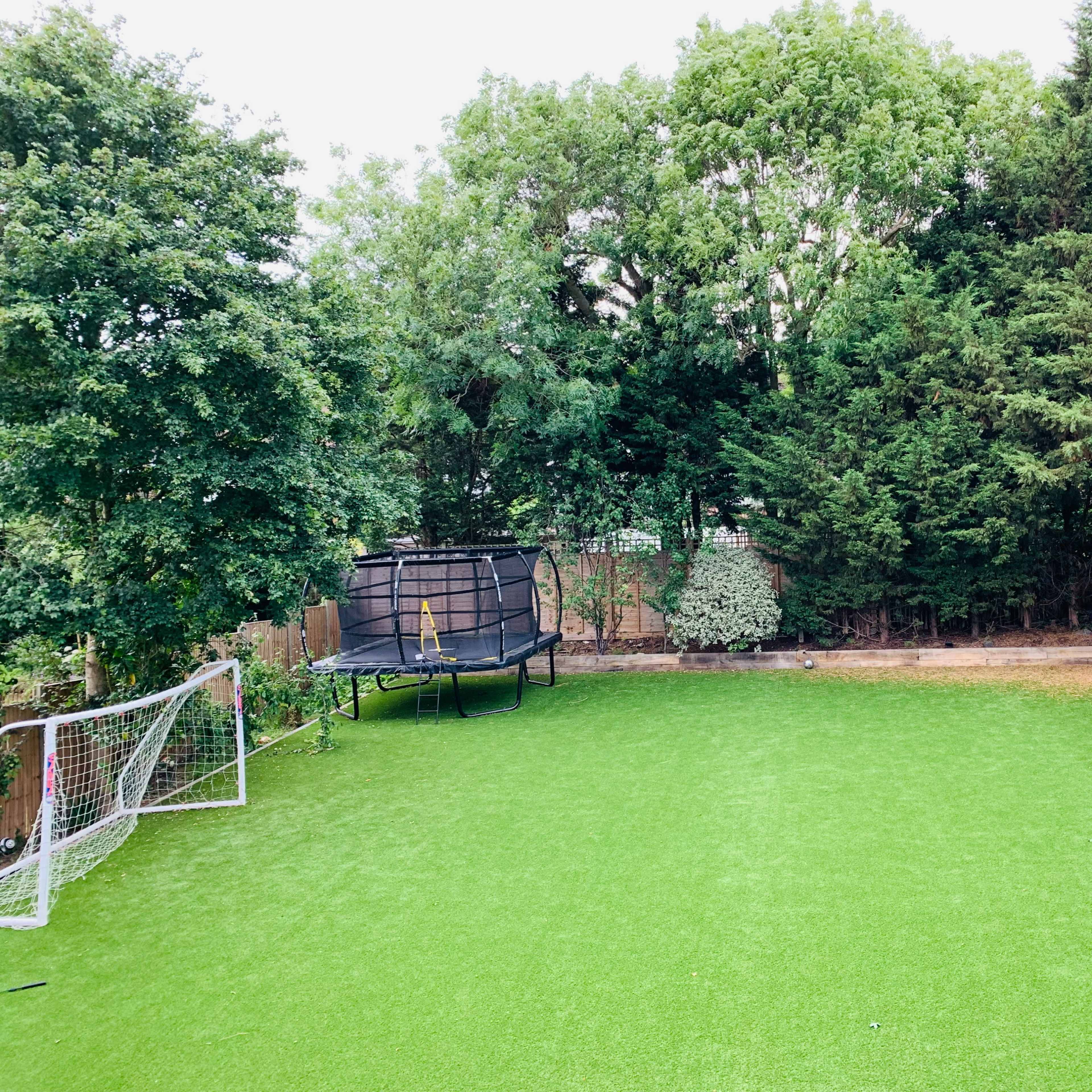 A neatly maintained artificial grass soccer field with two goalposts and a trampoline in a backyard surrounded by trees.