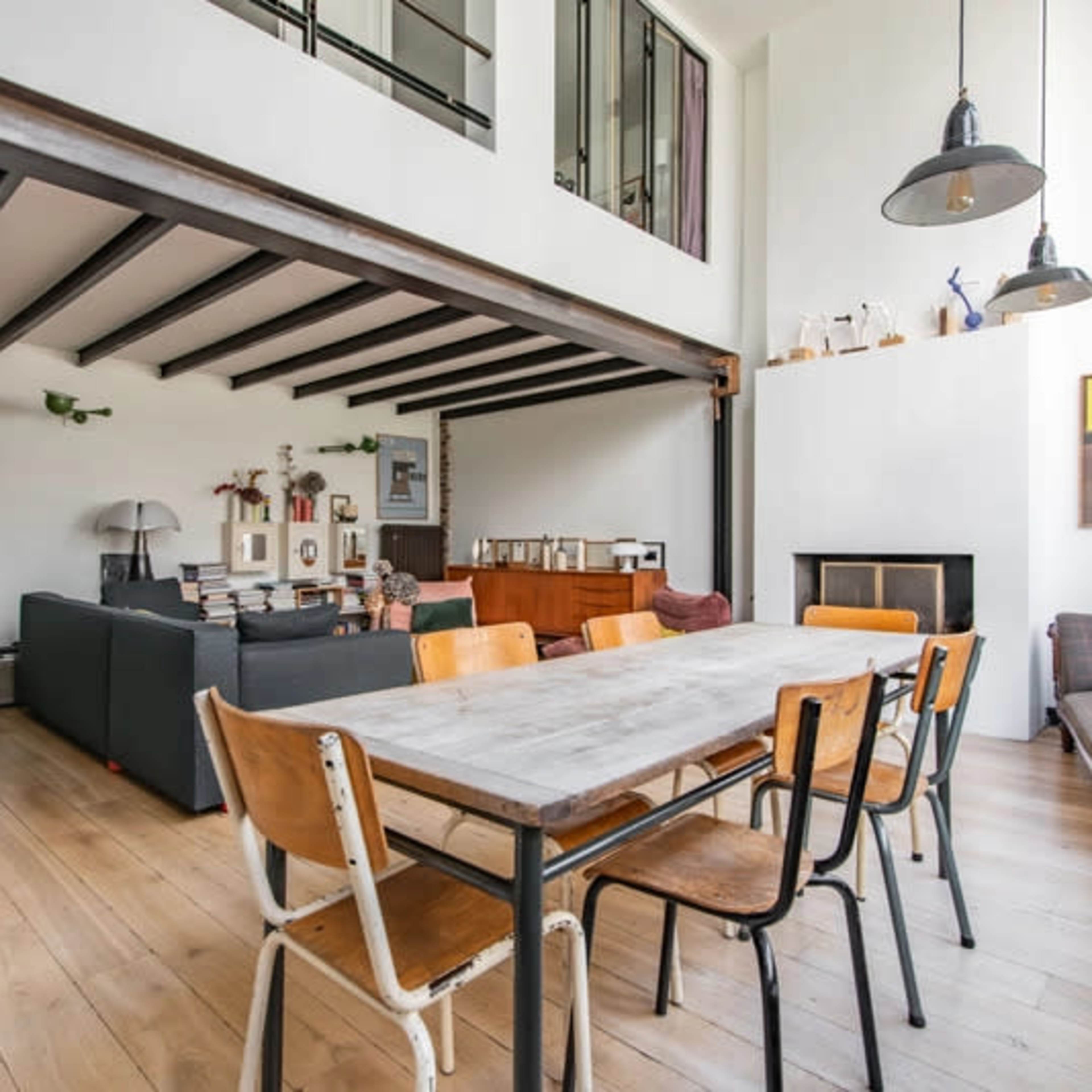 A spacious living area featuring a large wooden dining table surrounded by metal and wooden chairs, with a seating area visible in the background and a staircase leading to an upper level.
