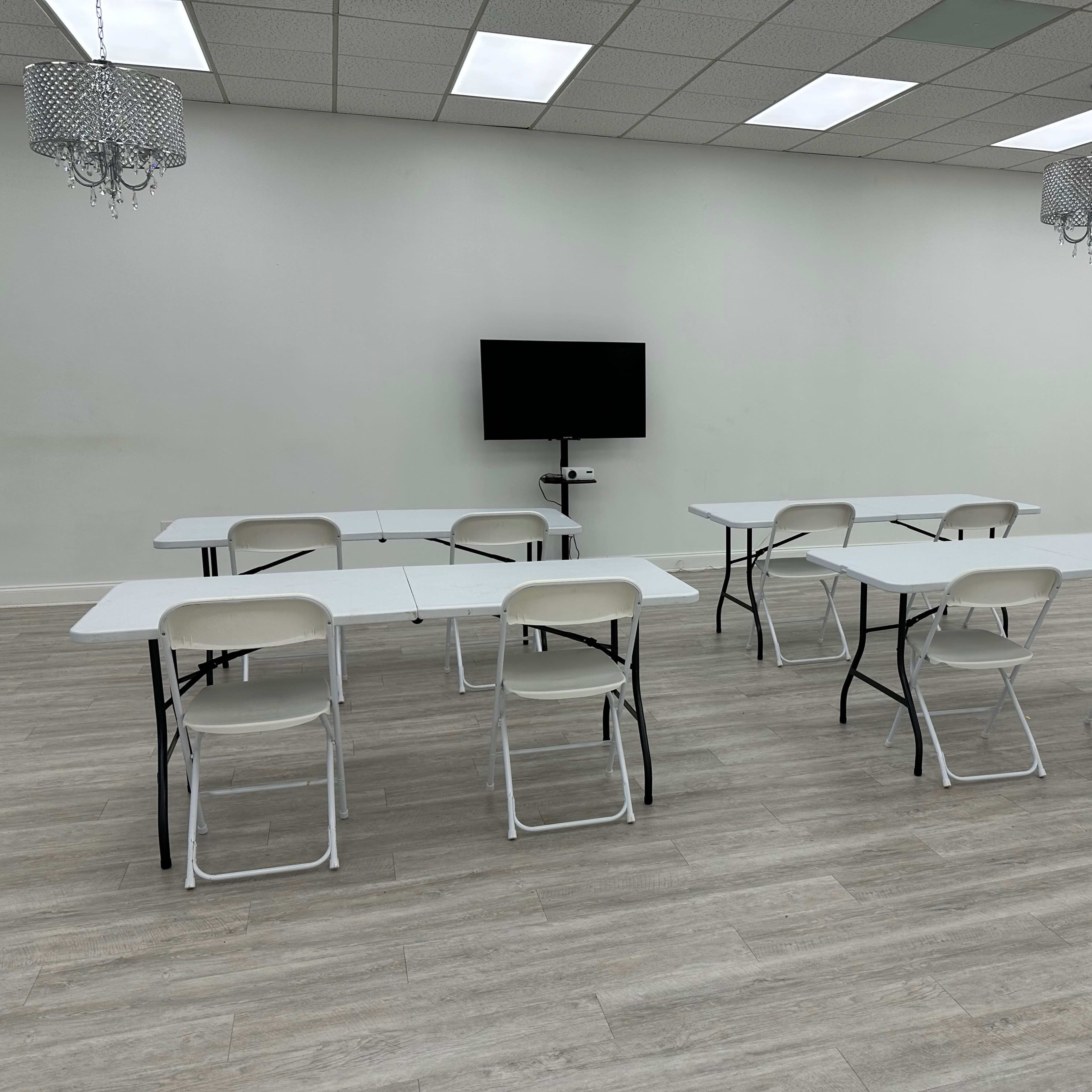 A simple, empty room features several folding tables and chairs arranged for a meeting, with a television mounted on the wall.