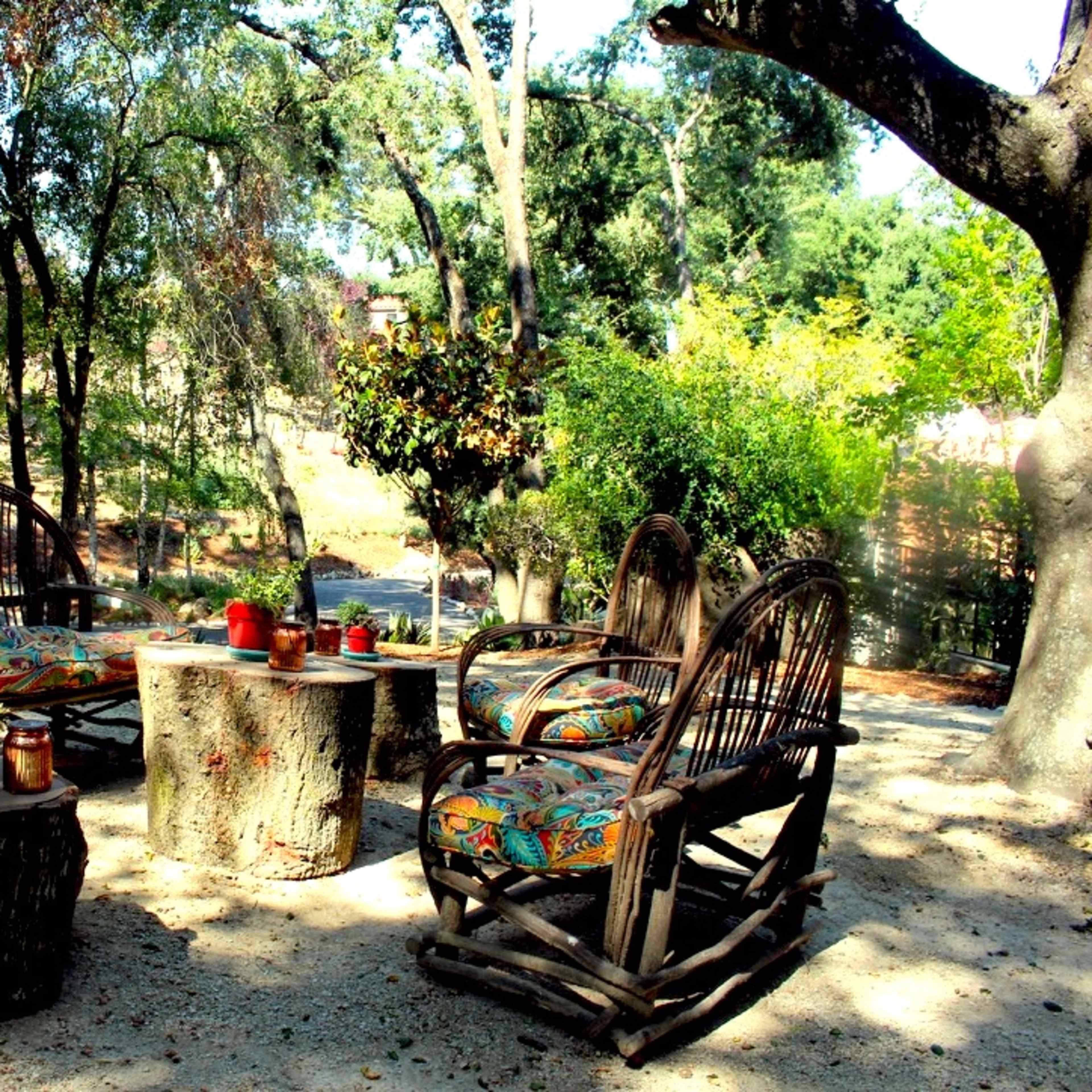 A seating area surrounded by trees features four chairs with colorful cushions arranged around a wooden stump table.