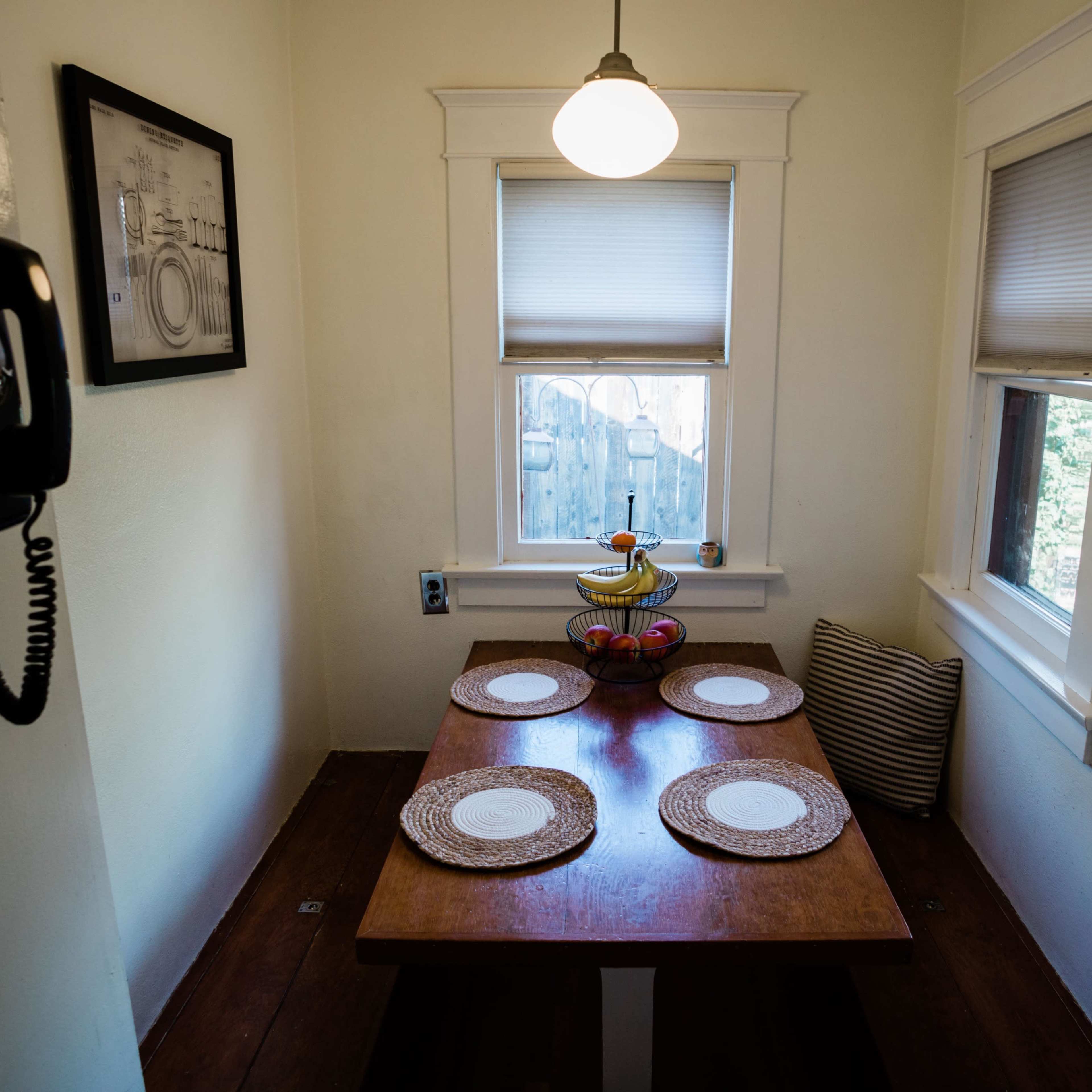 A small dining area features a wooden table set with four place settings and a fruit bowl, surrounded by windows with shades.