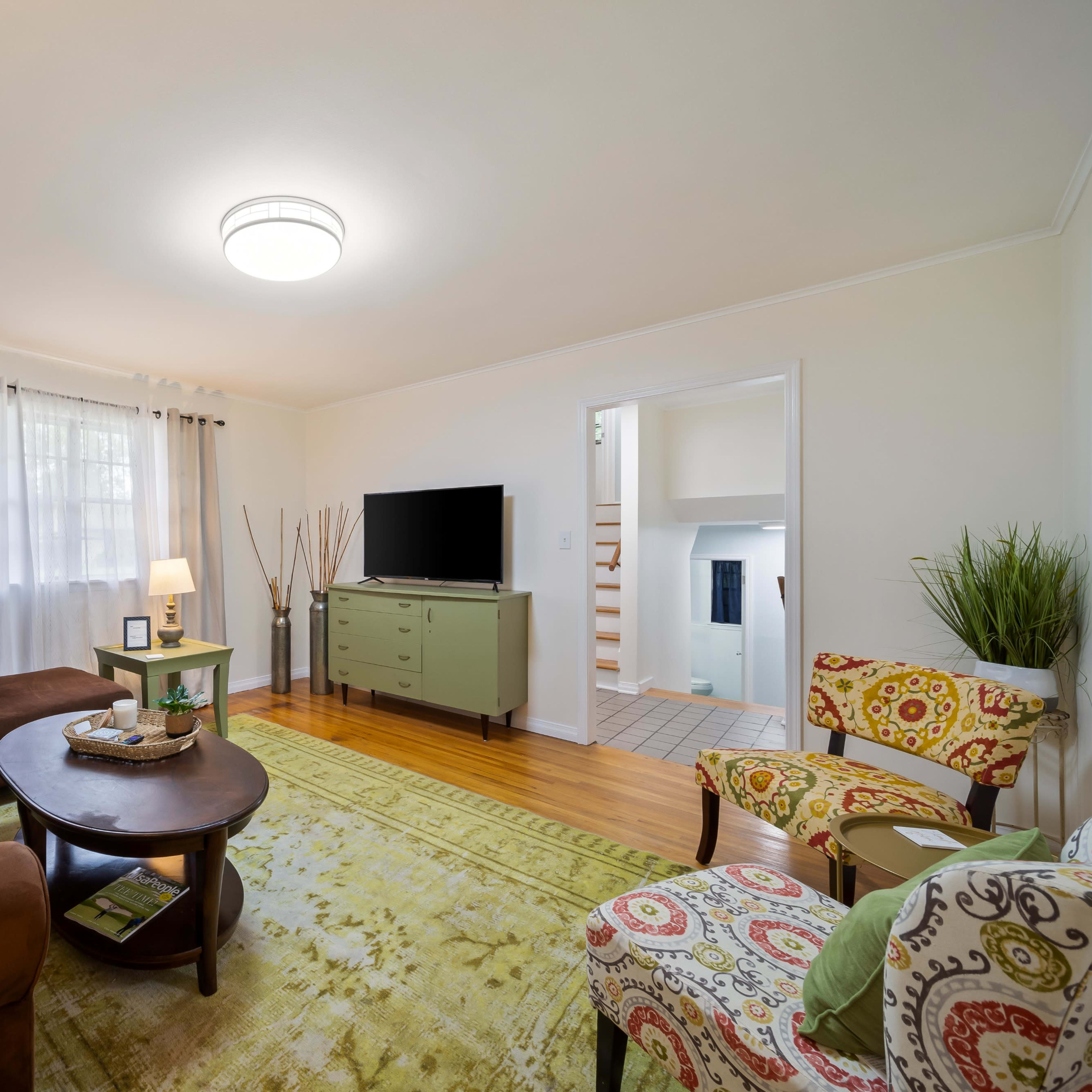 A cozy living room with a brown sectional sofa, a round coffee table, and colorful patterned chairs, featuring light-colored walls and a television on a green cabinet.