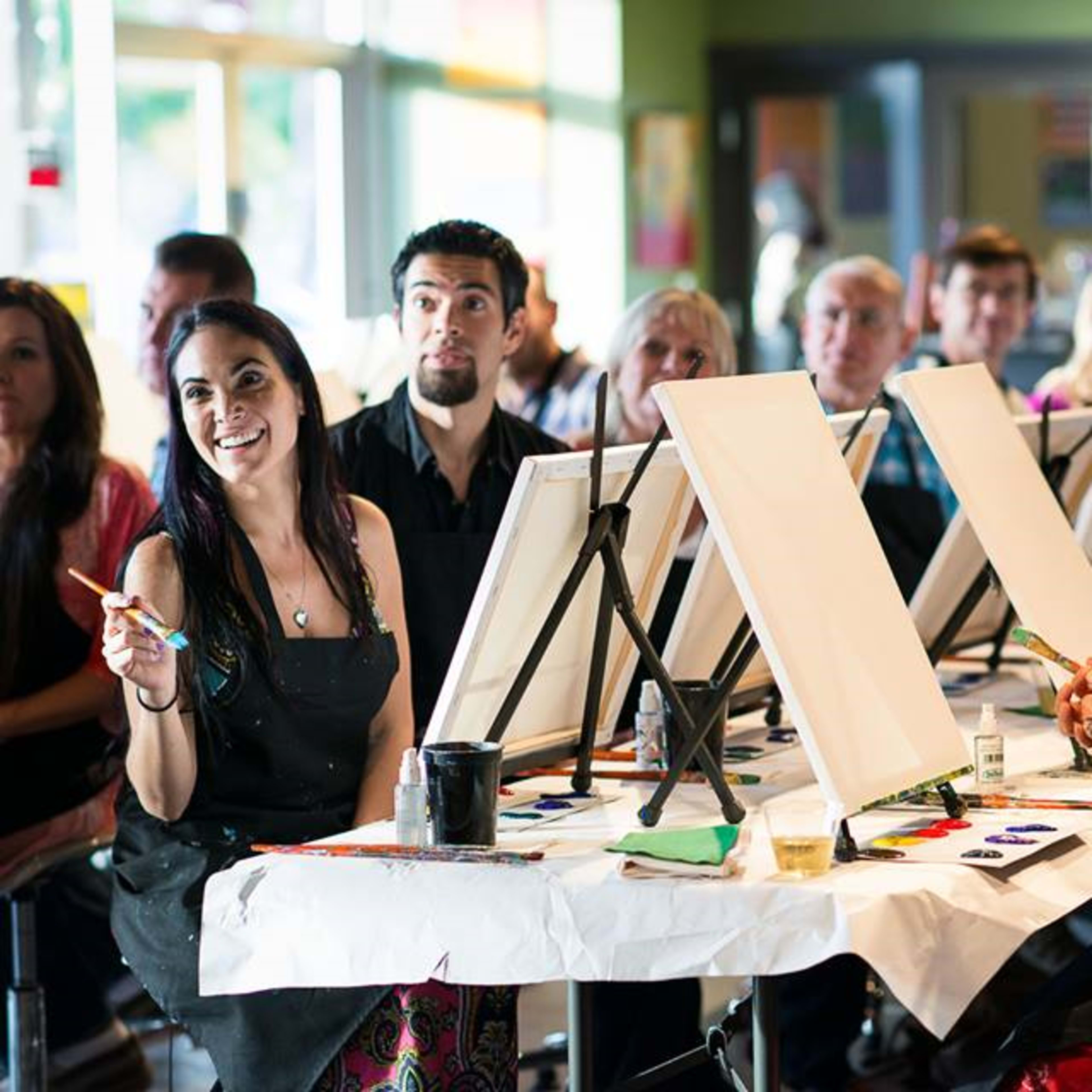 A group of people sit at tables in an art class, facing blank canvases as two women hold paintbrushes.