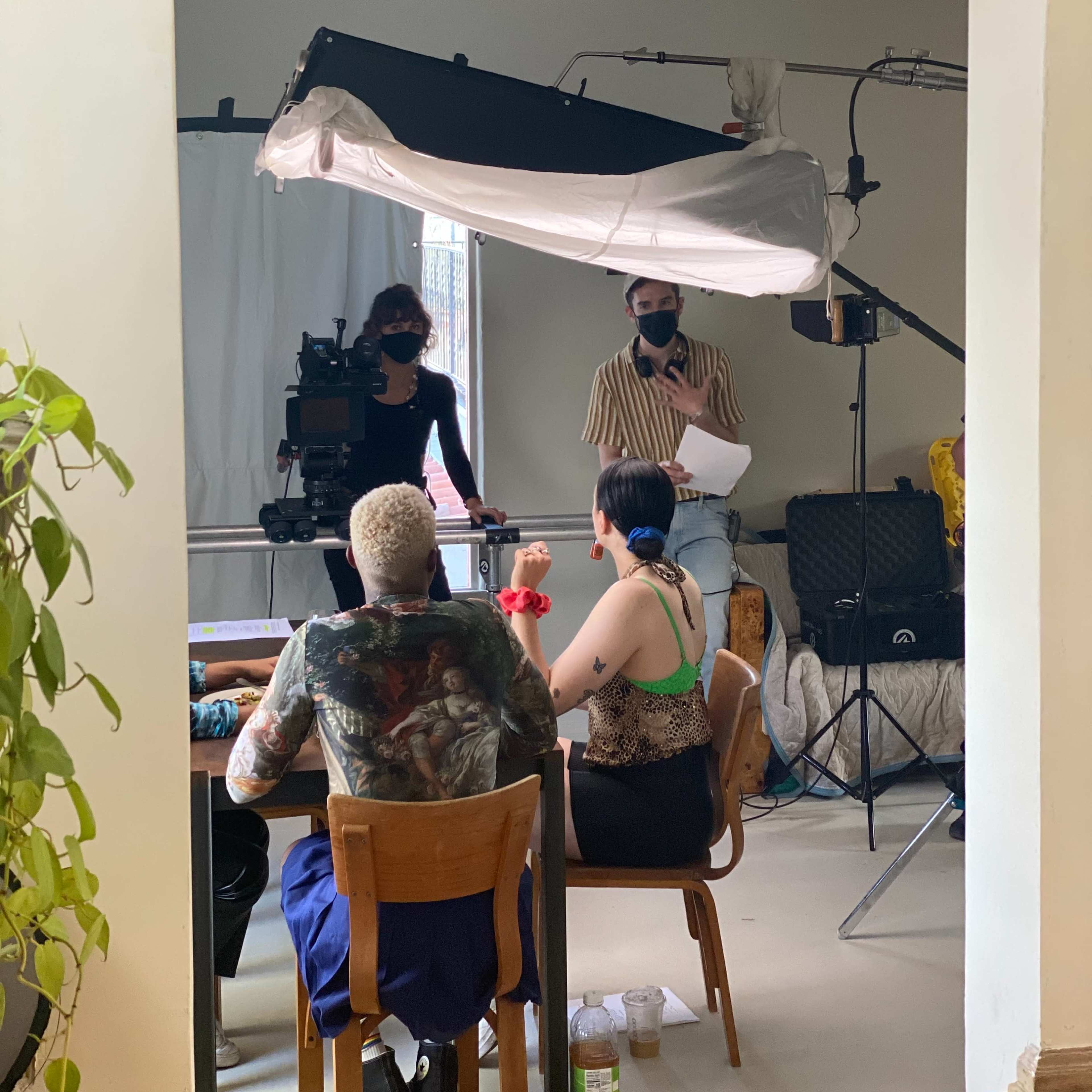 A film crew is set up in a room with two people seated at a table while a woman stands and directs, with lighting and camera equipment positioned nearby.