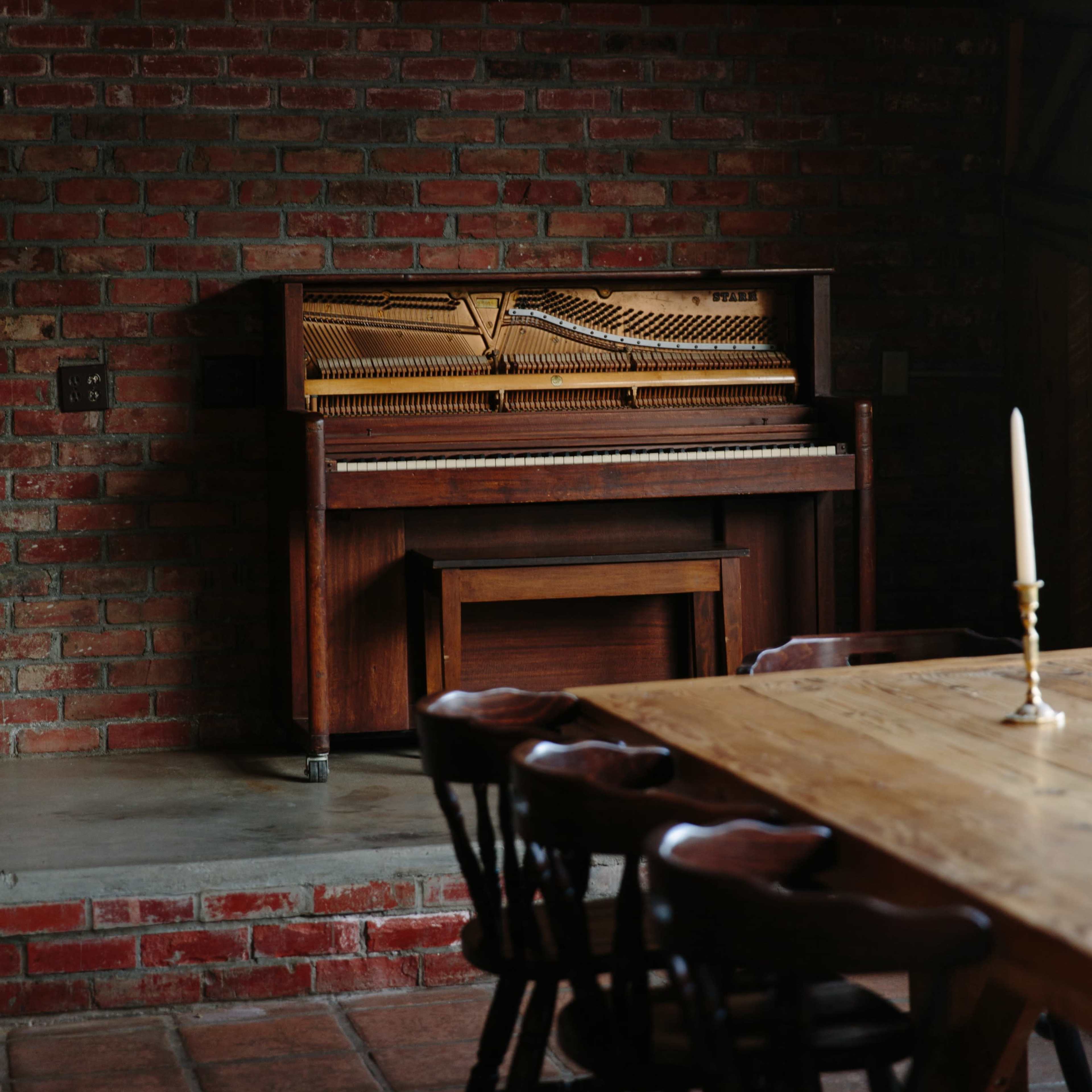 A wooden piano sits against a brick wall next to a long wooden dining table with chairs.