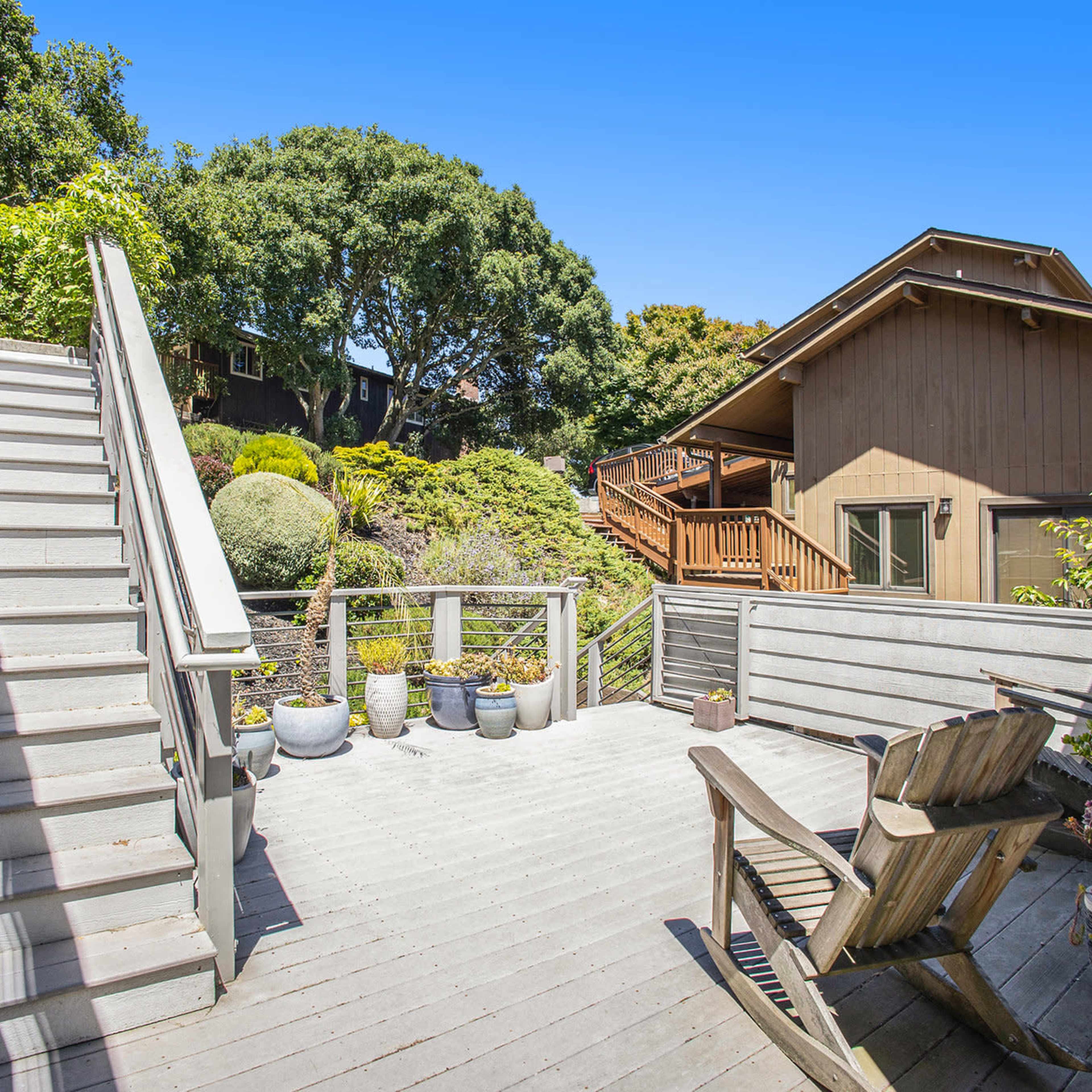 A wooden deck with stairs leads to a landscaped backyard featuring potted plants and a neighboring house.