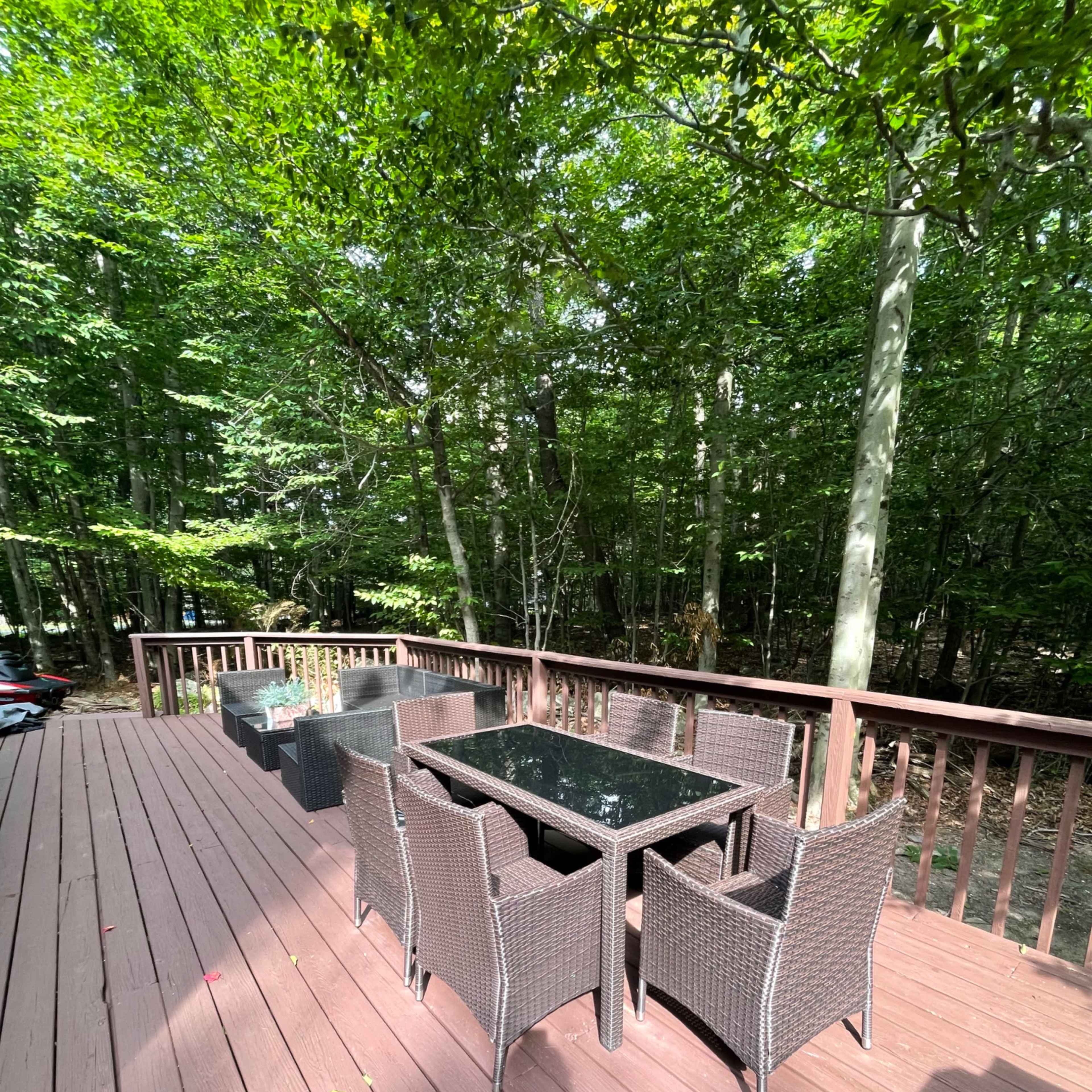 A wooden deck surrounded by trees features a glass-top dining table with eight wicker chairs.