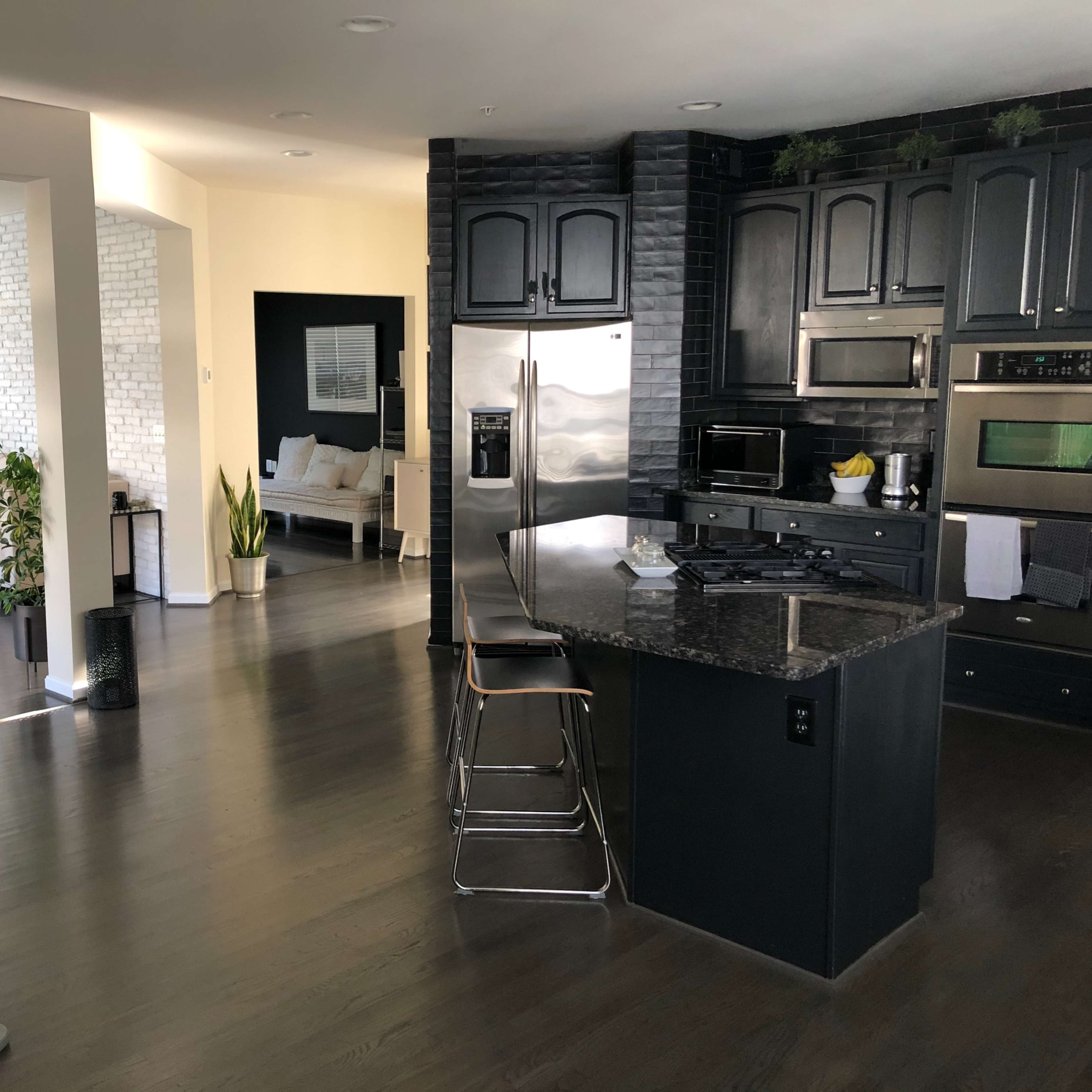 A modern kitchen with dark cabinetry, a granite countertop, and a view into a light-filled living area featuring a sofa and plants.