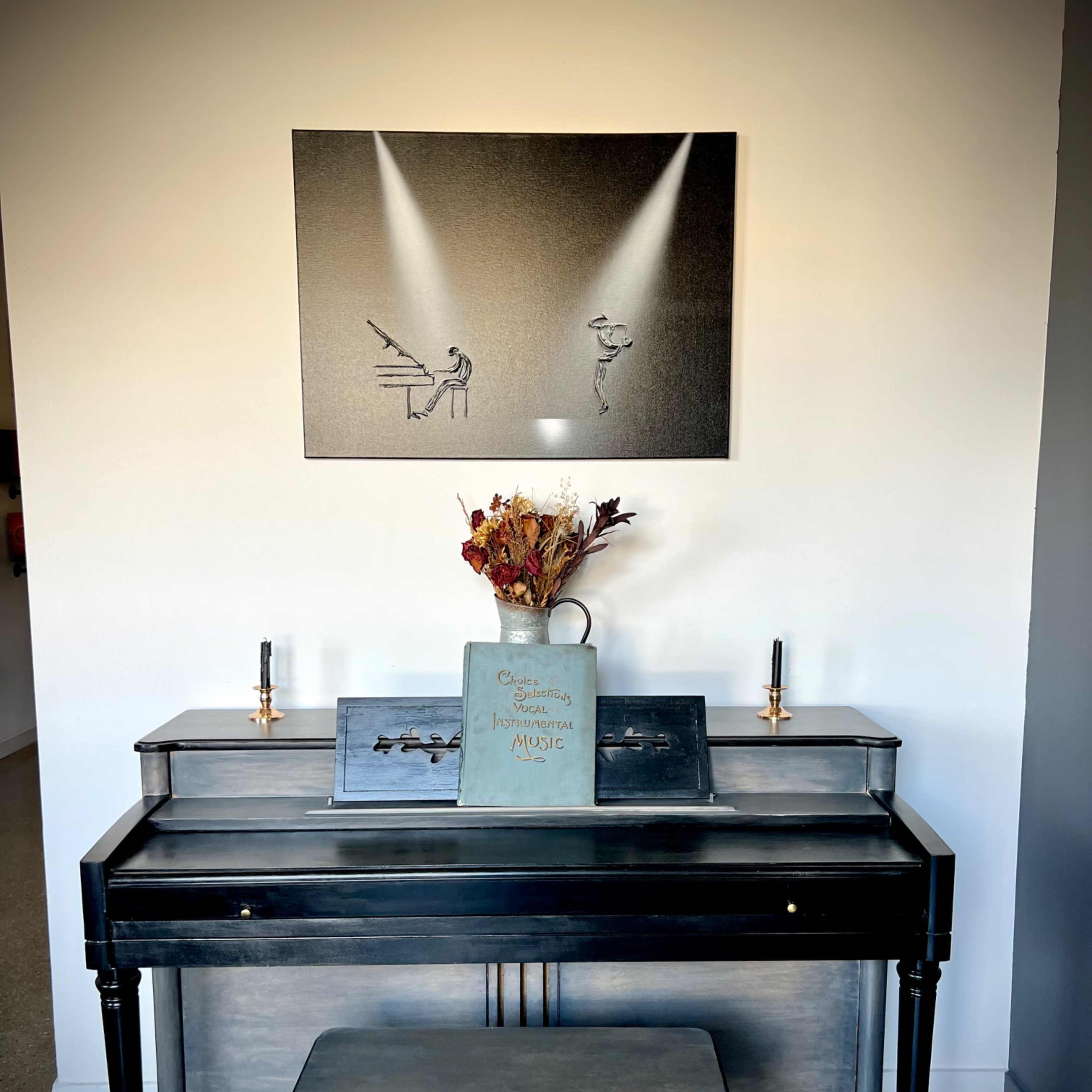 A vintage piano with a bench sits against a gray wall, accompanied by a framed artwork depicting a musician and a dancer, and a vase with dried flowers on top.
