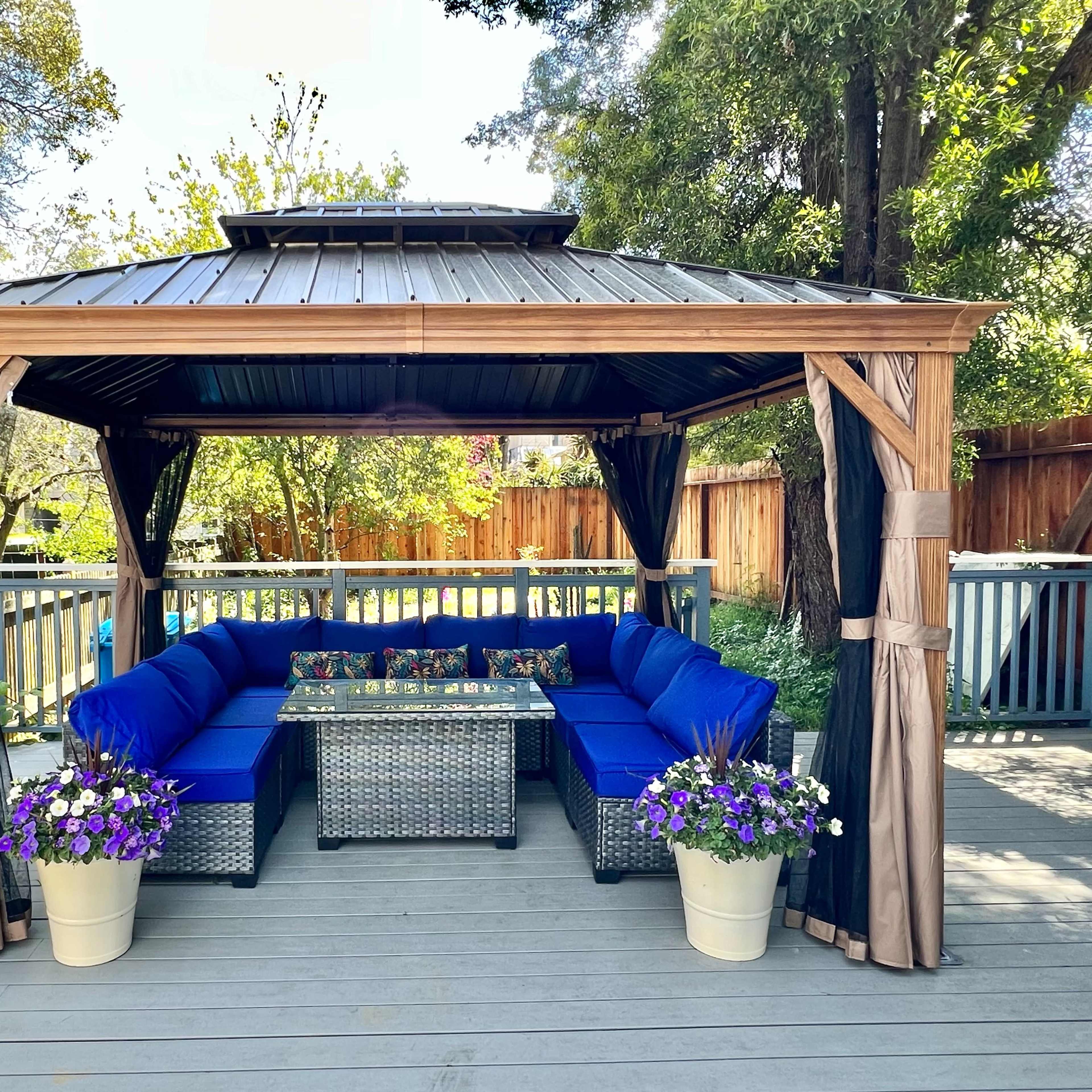 A gazebo with blue upholstered seating surrounds a glass-top table, placed on a deck adorned with potted purple flowers.