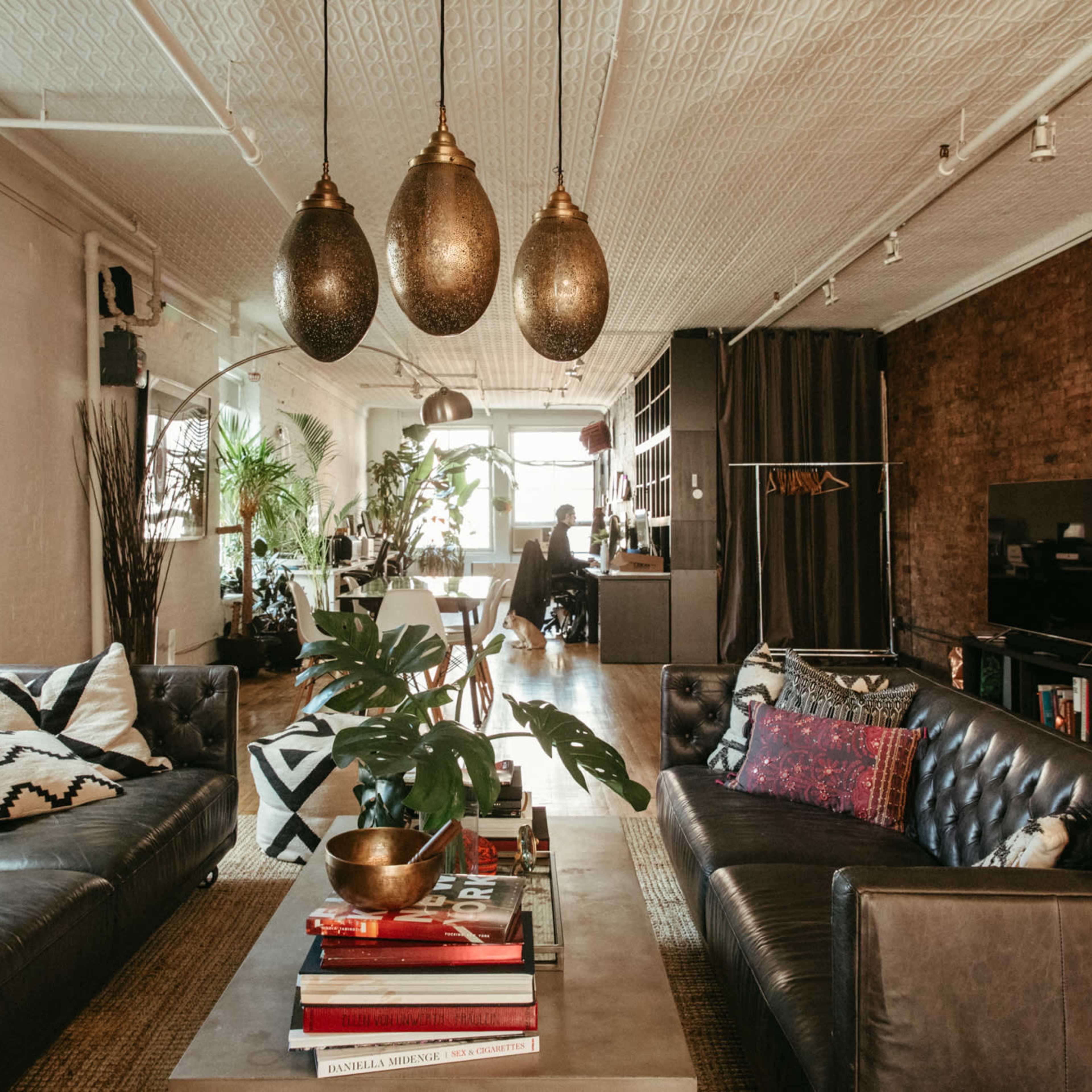 A stylish living room with a dark leather sofa, a central coffee table stacked with books, and large pendant lights hanging from the ceiling, alongside greenery and a modern TV setup against a brick wall.