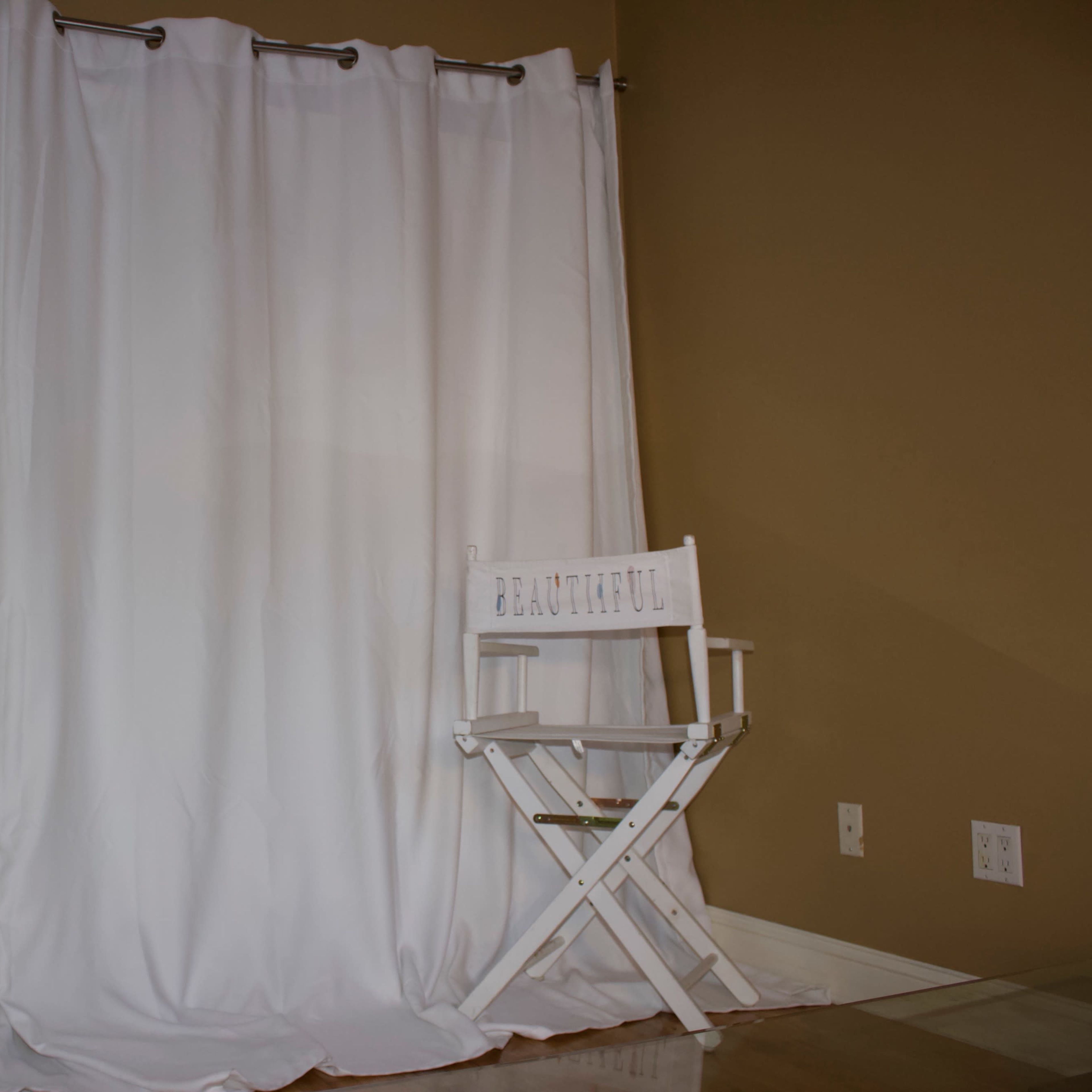 A white chair labeled "BEAUTIFUL" sits in front of a set of white curtains against a beige wall.