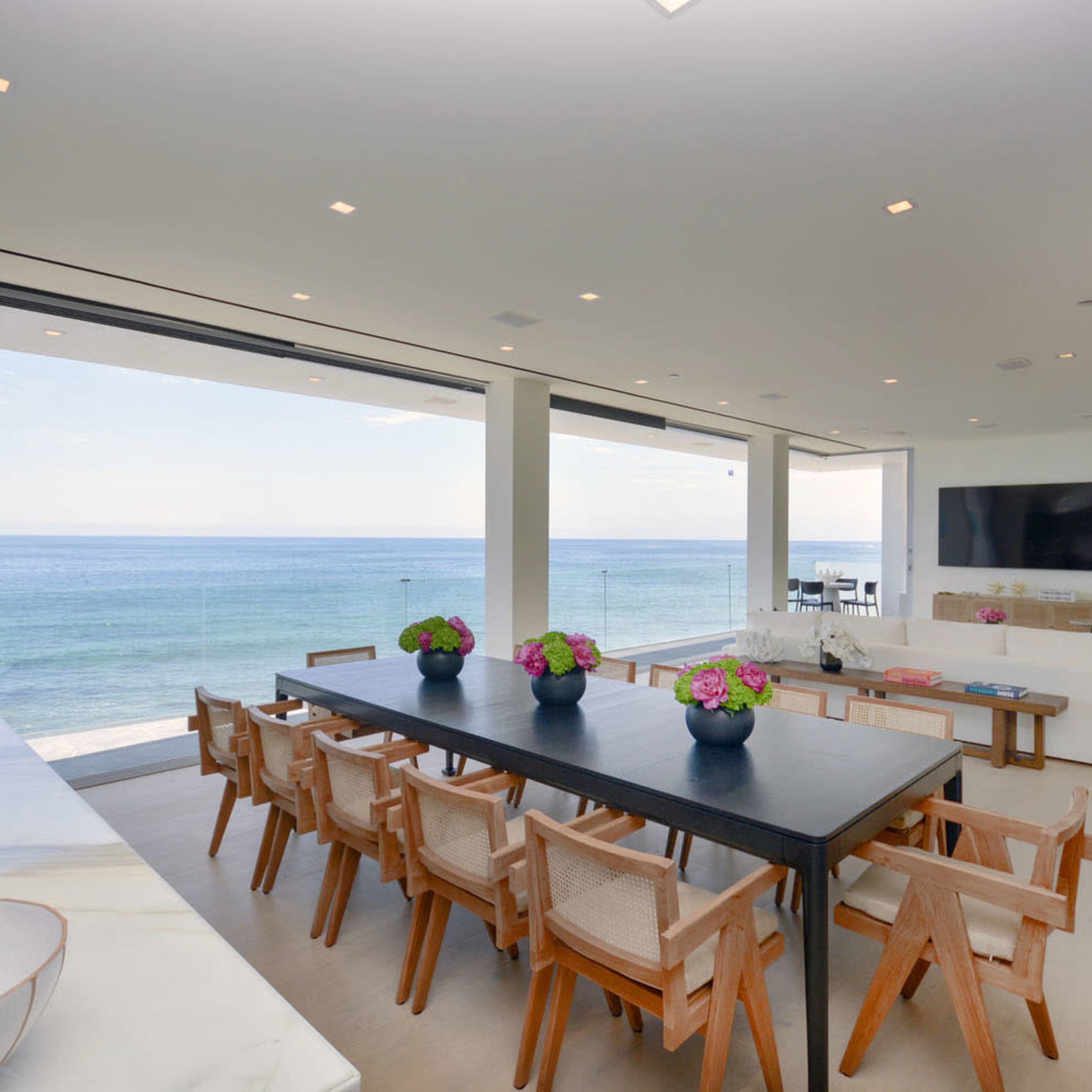 A modern dining area features a long black table surrounded by wooden chairs, with large windows offering a view of the ocean.
