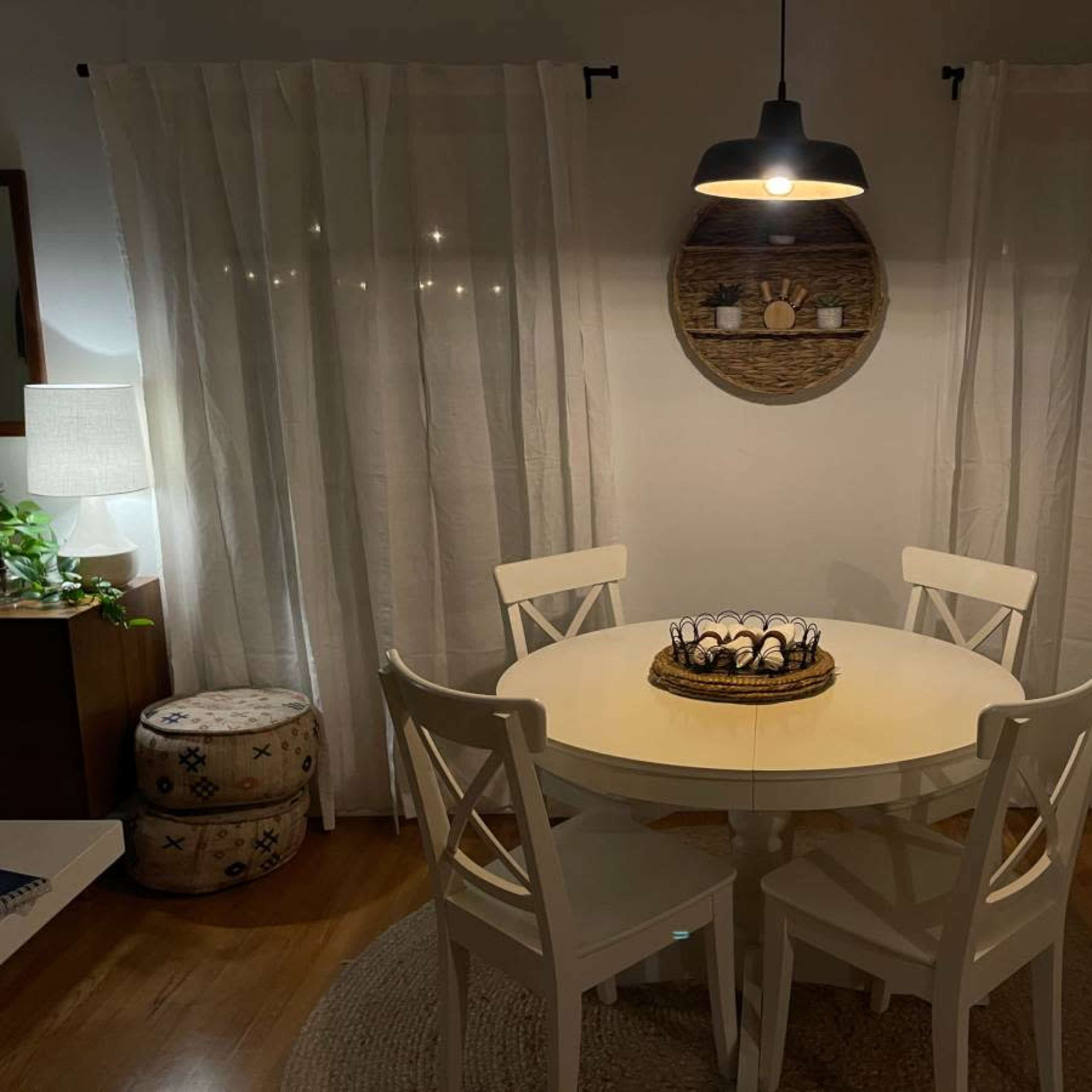 A round white dining table with four chairs is centered in a softly lit room, surrounded by light curtains and a woven wall decor.