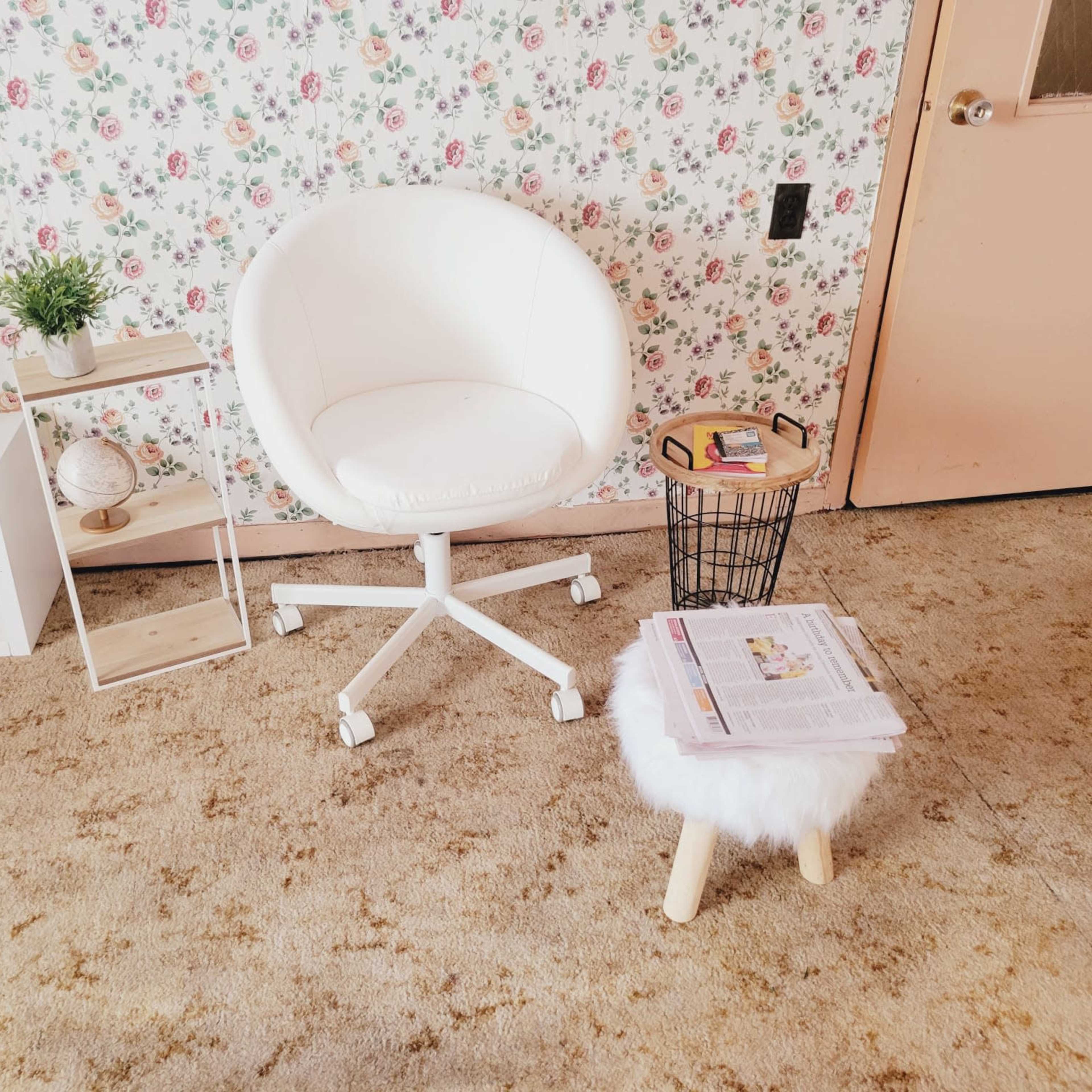 A small living space with a white swivel chair, a round stool with a fluffy seat, a small side table holding a magazine, and a decorative shelf against a floral wallpaper background.