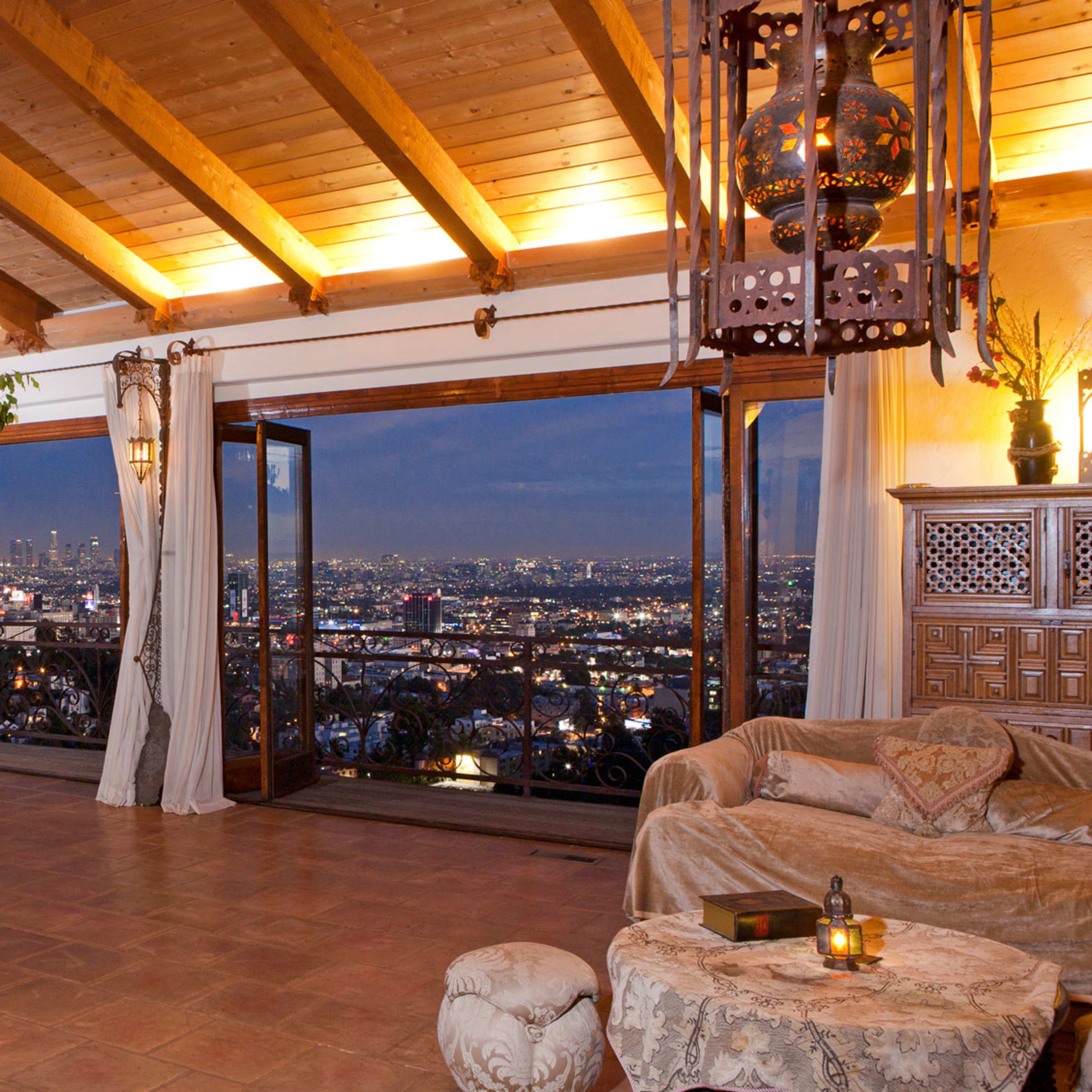 A spacious living room features large windows overlooking a cityscape, with wooden beams, comfortable seating, and decorative plants.