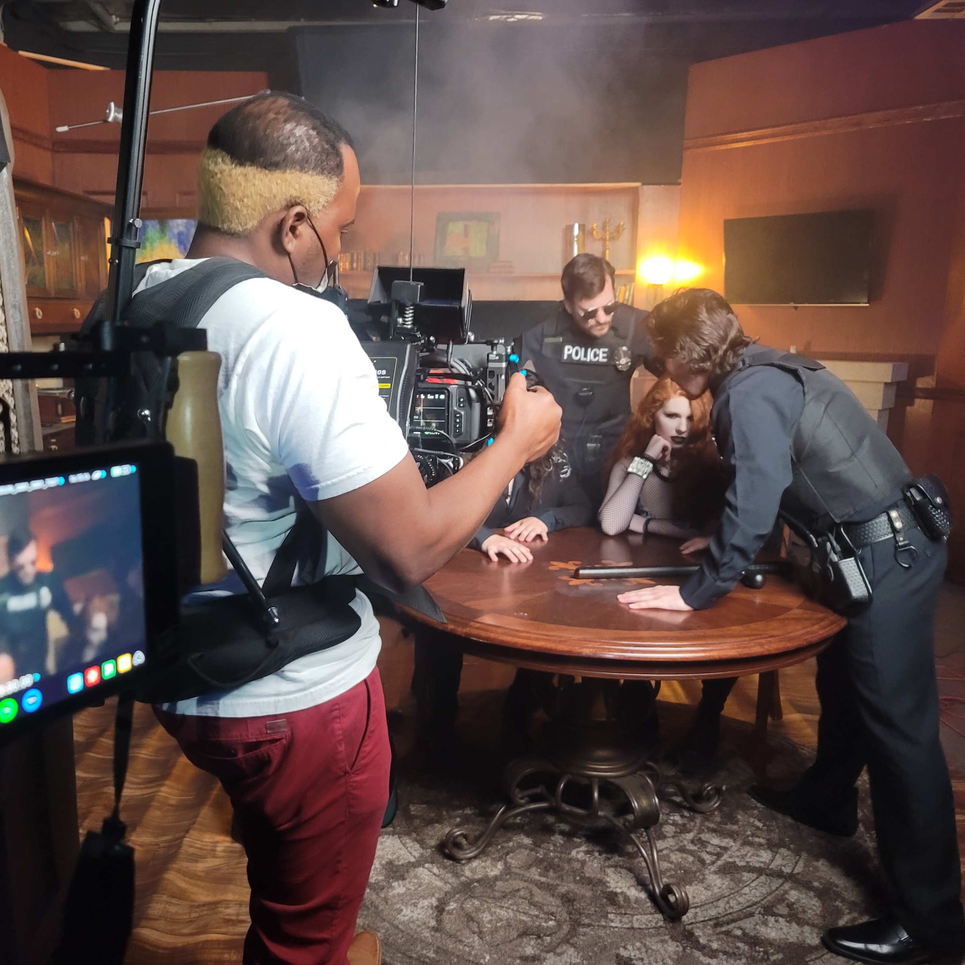 A film crew captures a scene with police officers and a suspect gathered around a table in a dimly lit room.