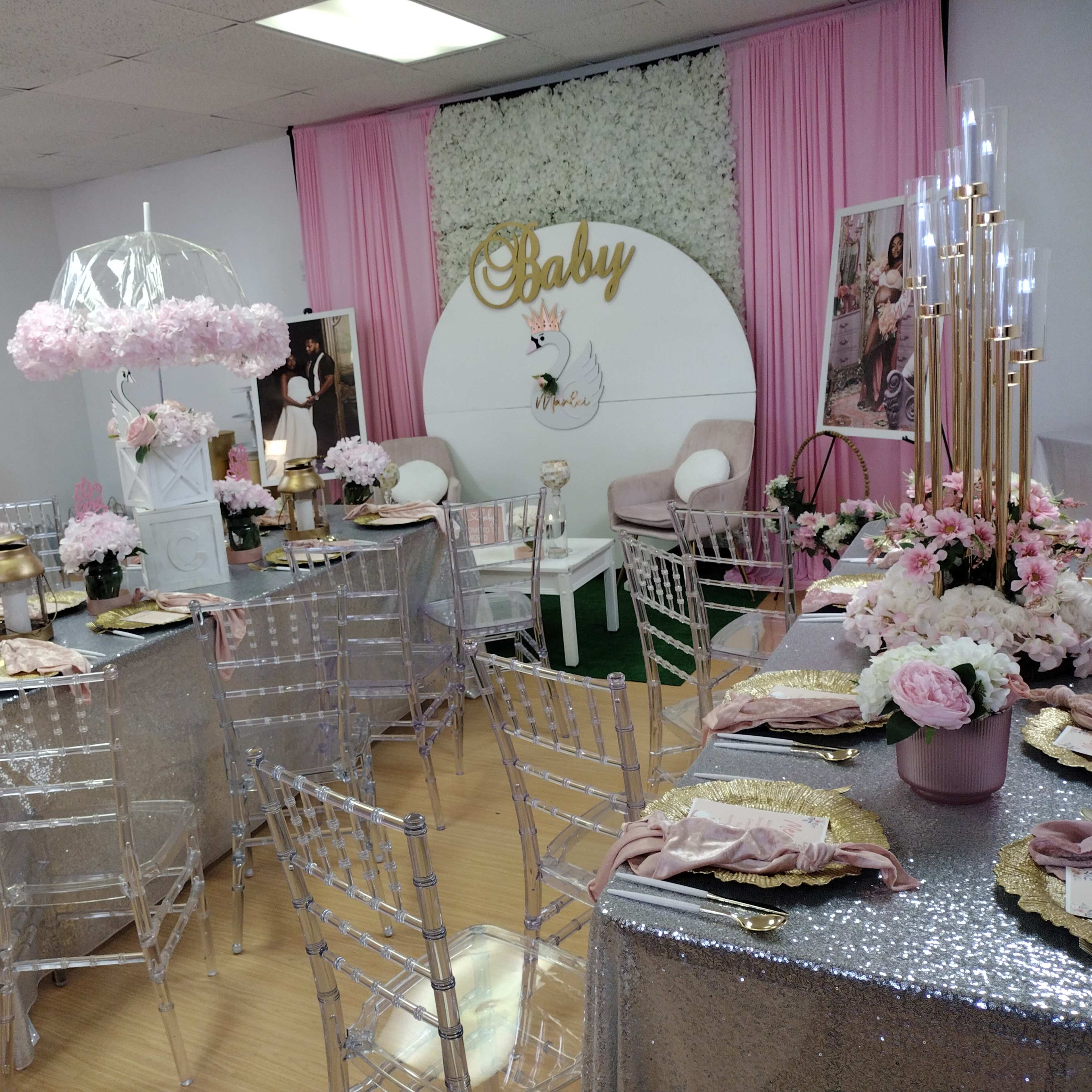 A decorated room for a baby shower, featuring tables covered in silver tablecloths, floral centerpieces, and a backdrop with the word "Baby."