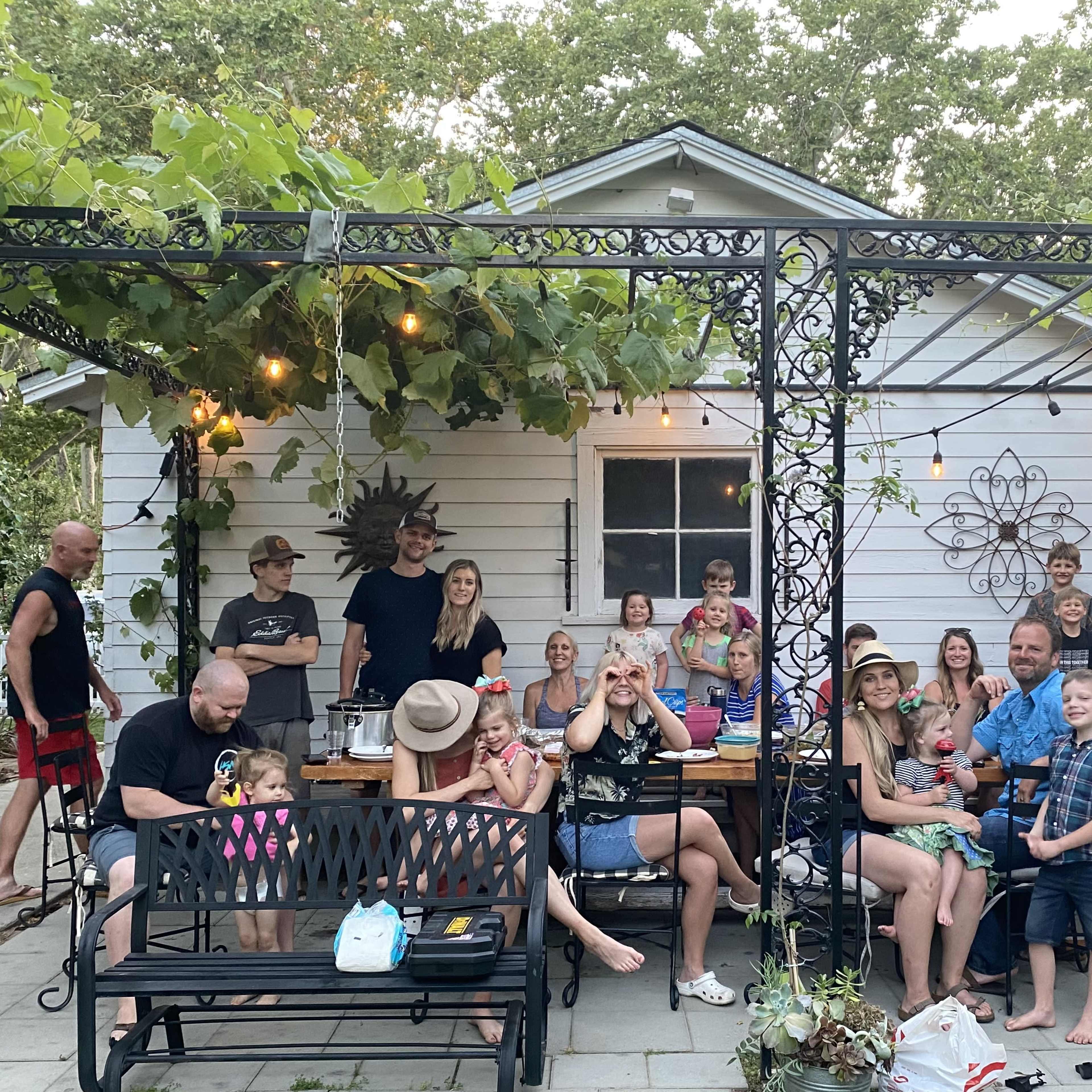 A large group of people, including children and adults, gather on a patio decorated with string lights and surrounded by greenery.