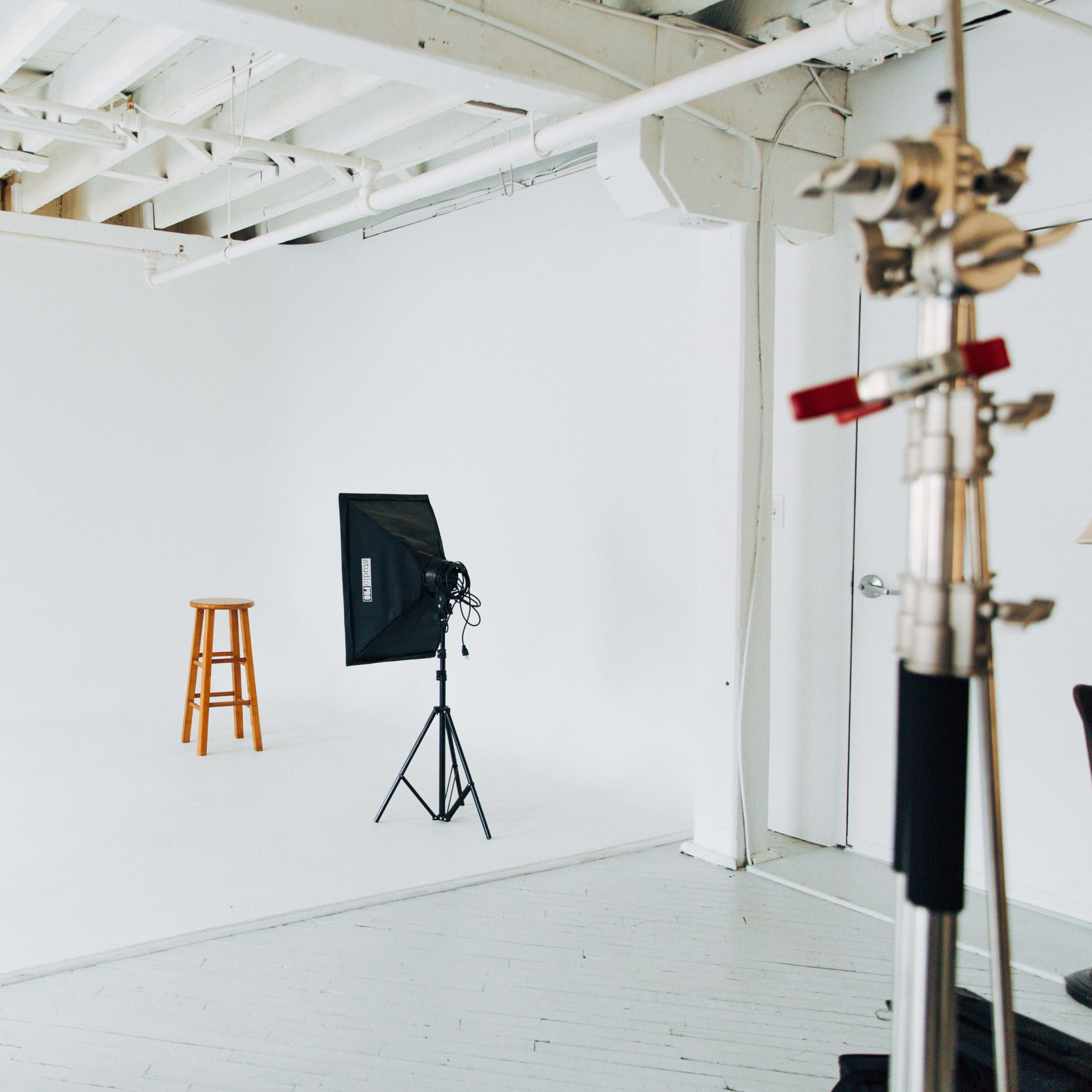 A photography studio features a white backdrop, a wooden stool in the center, and two softbox lights on stands.