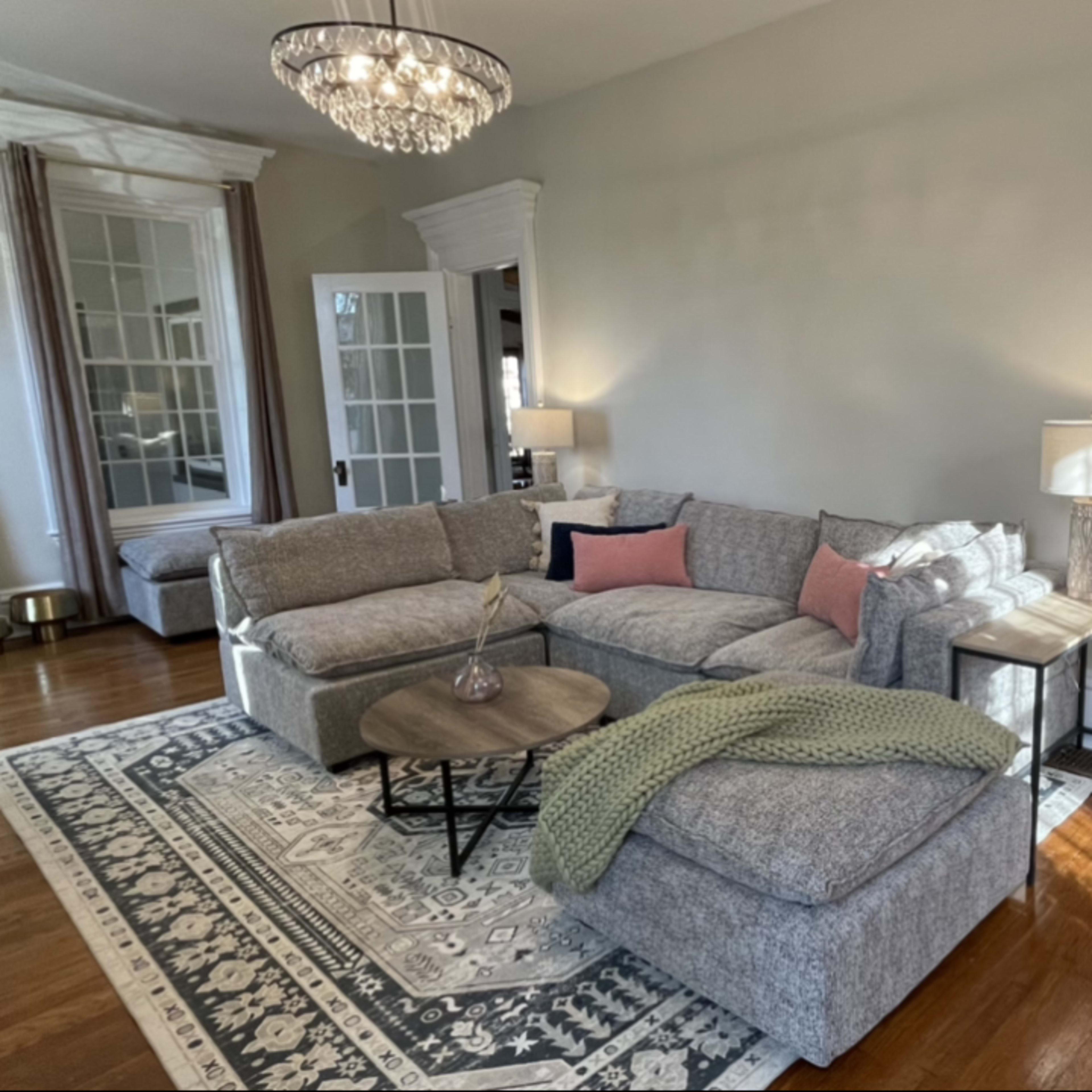 A well-lit living room featuring a grey sectional sofa, a round coffee table, and a large mirror, with French doors leading to a bright outdoor area.