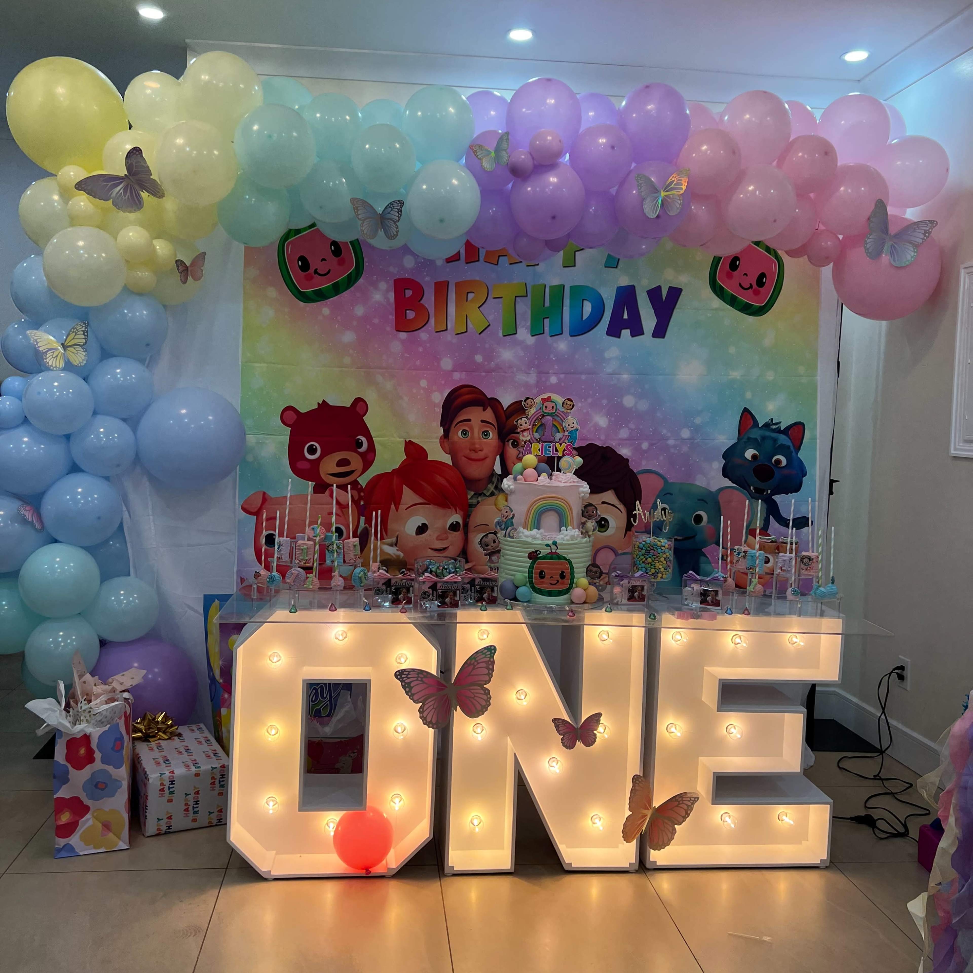 A colorful birthday celebration setup featuring a "ONE" light-up sign, balloons, and a decorated table with a cake and themed decorations in the background.