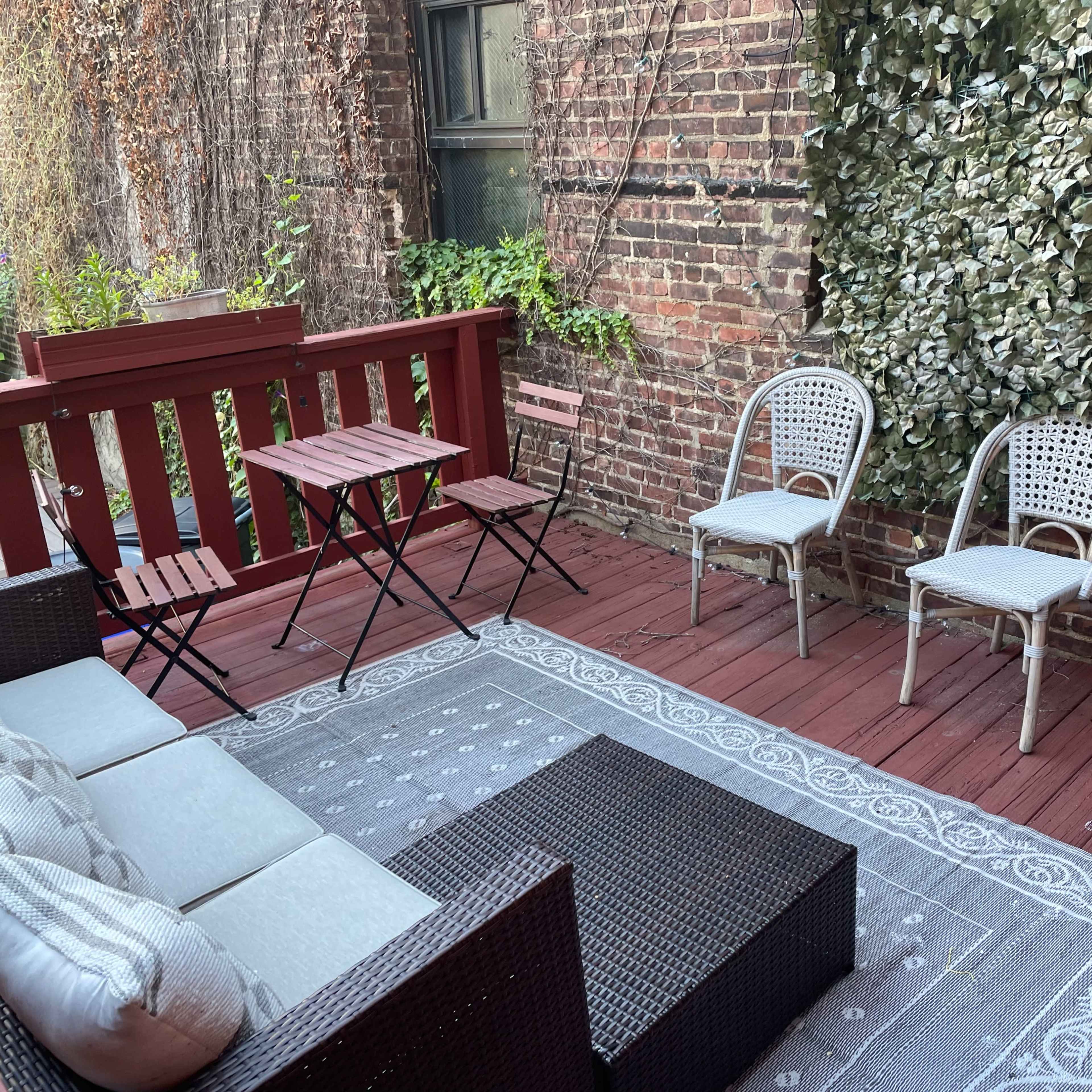 A small outdoor deck area with a seating arrangement, a low coffee table, two chairs, and decorative elements against a brick wall.